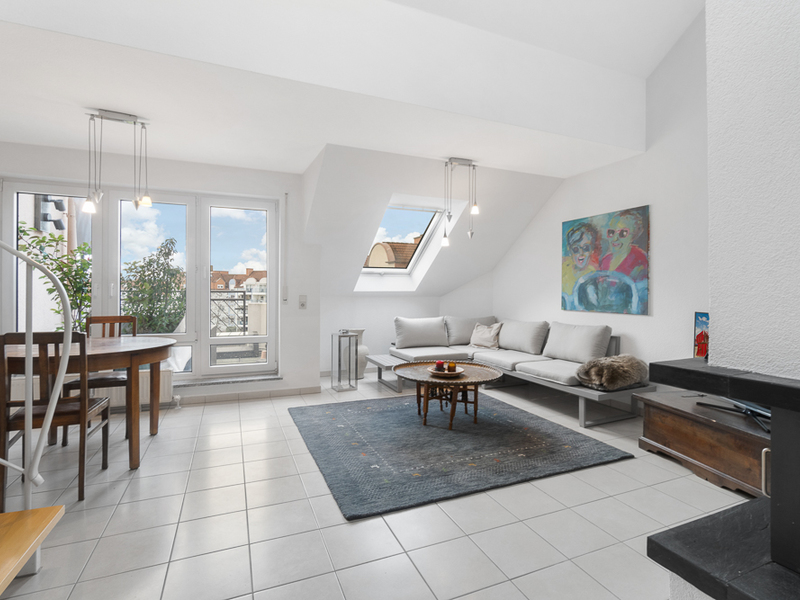 Helles Wohnzimmer mit Sofa, Esstisch, blauem Teppich und großen Fenstern mit Blick nach draußen.