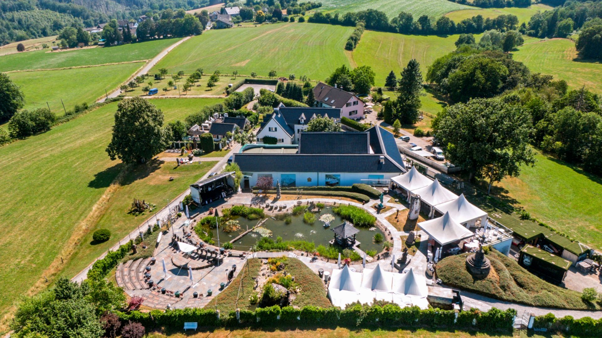 Luftaufnahme eines Wohnhauses mit Garten, Teich und weißen Pavillons, umgeben von Wiesen und Bäumen.