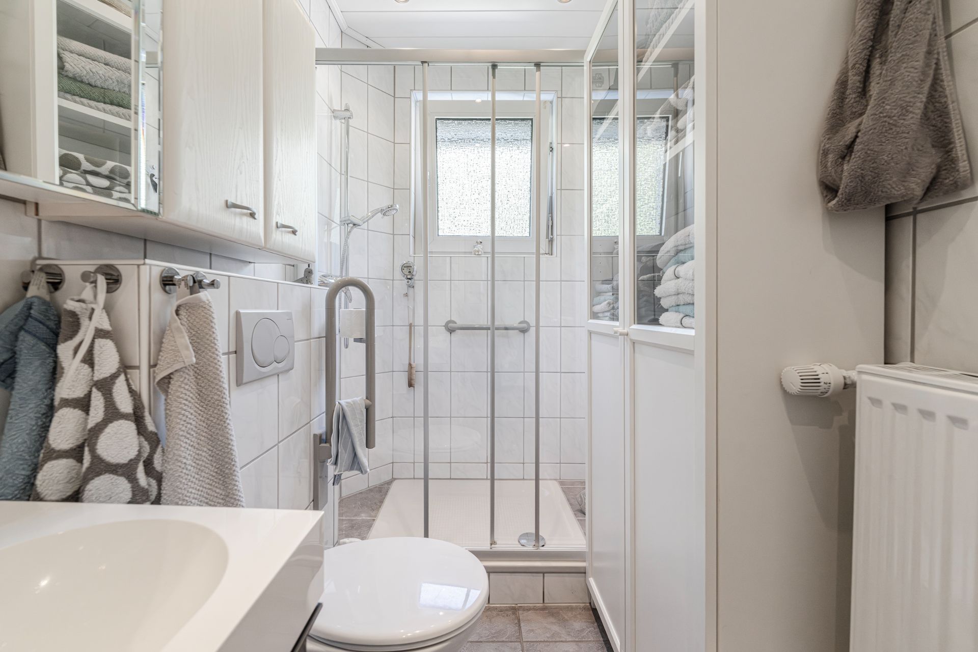 Modernes, kleines Badezimmer mit Dusche, WC, Waschbecken, Handtüchern und weißen Schränken.
