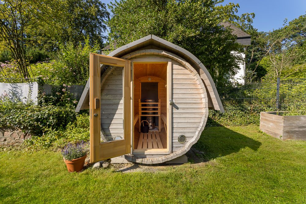 Runde Holzsauna im Garten mit geöffneter Glastür, umgeben von Gras und Pflanzen.