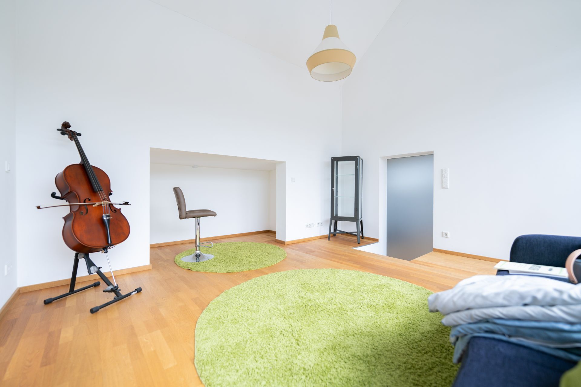 Helles Zimmer mit Holzfußboden, grünem Teppich, Cello, Stuhl und Sofa.