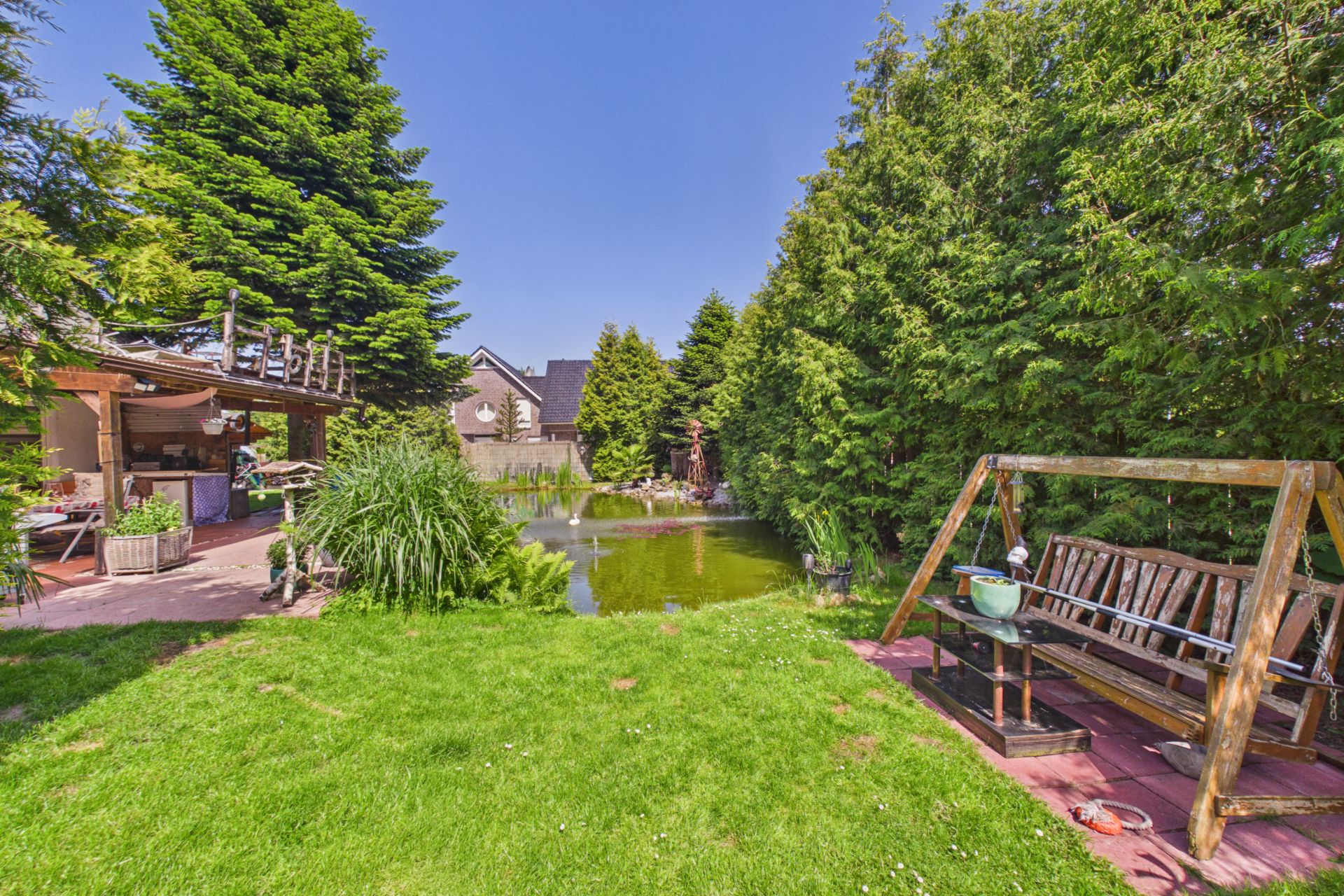 Garten mit Holzschaukel, Teich, Rasenfläche und großen Bäumen.