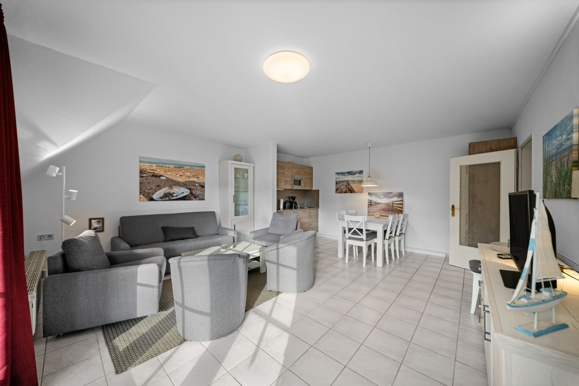 Helles Wohnzimmer mit grauem Sofa, zwei Sesseln, Esstisch, Fernseher und Wandbildern.