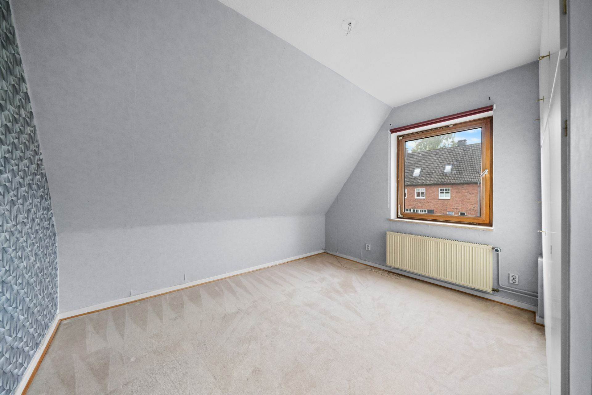 Leeres Zimmer mit schräger Decke, großem Fenster, Heizkörper und heller Teppichboden.