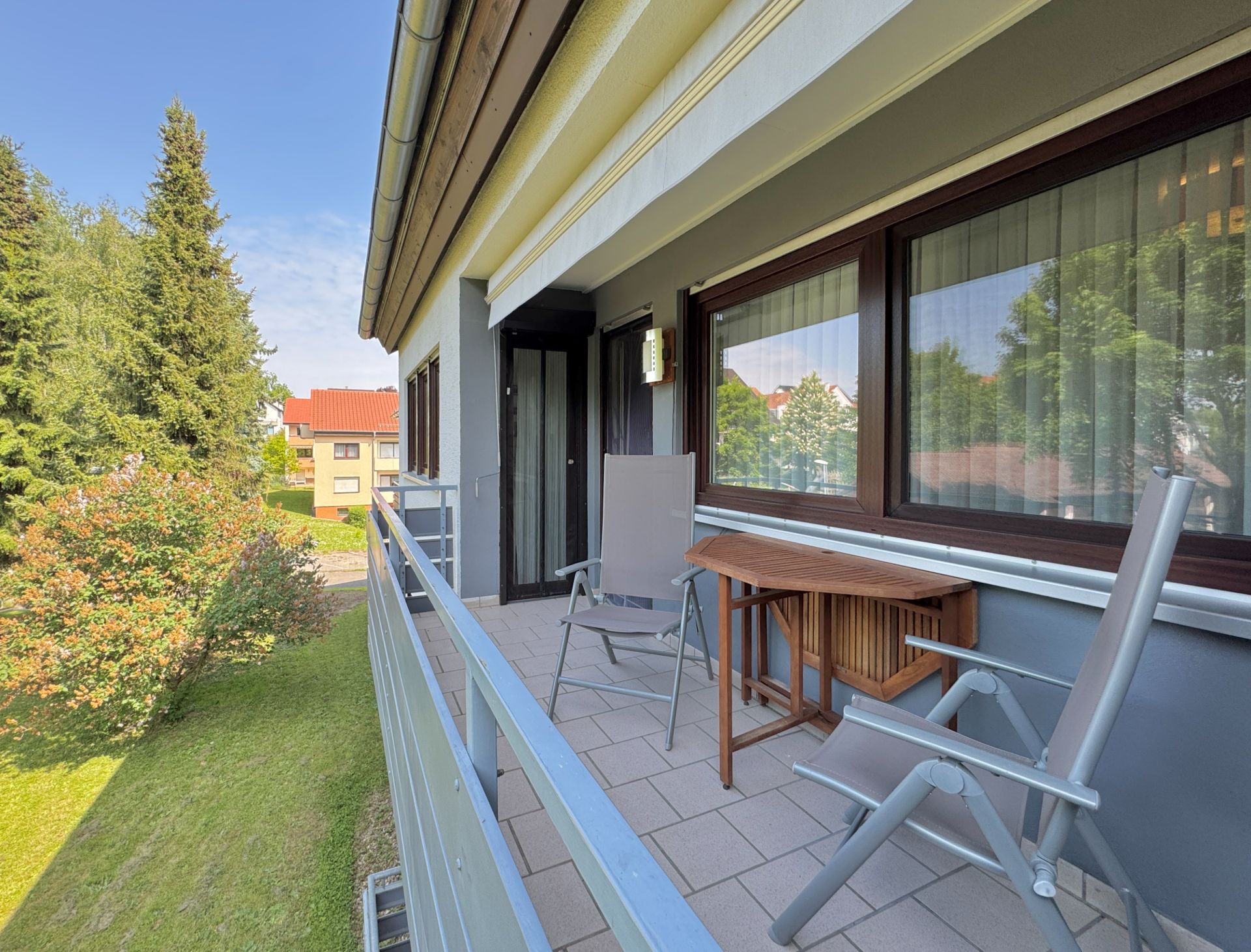 Balkon mit zwei Stühlen und einem kleinen Holztisch, Blick auf Garten und Bäume.