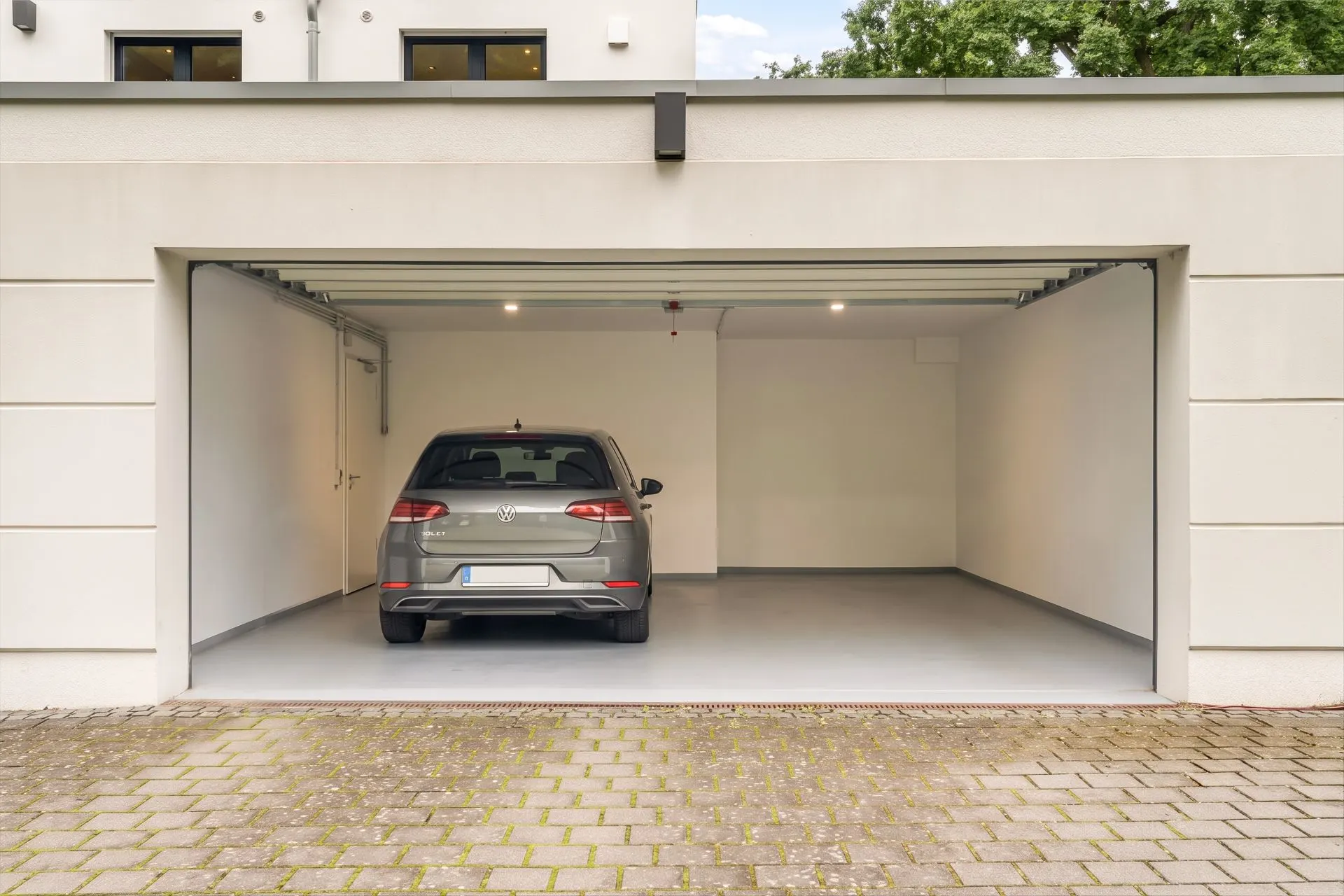Offene Garage mit einem einzelnen Auto darin, weiße Wände und gepflasterte Einfahrt.