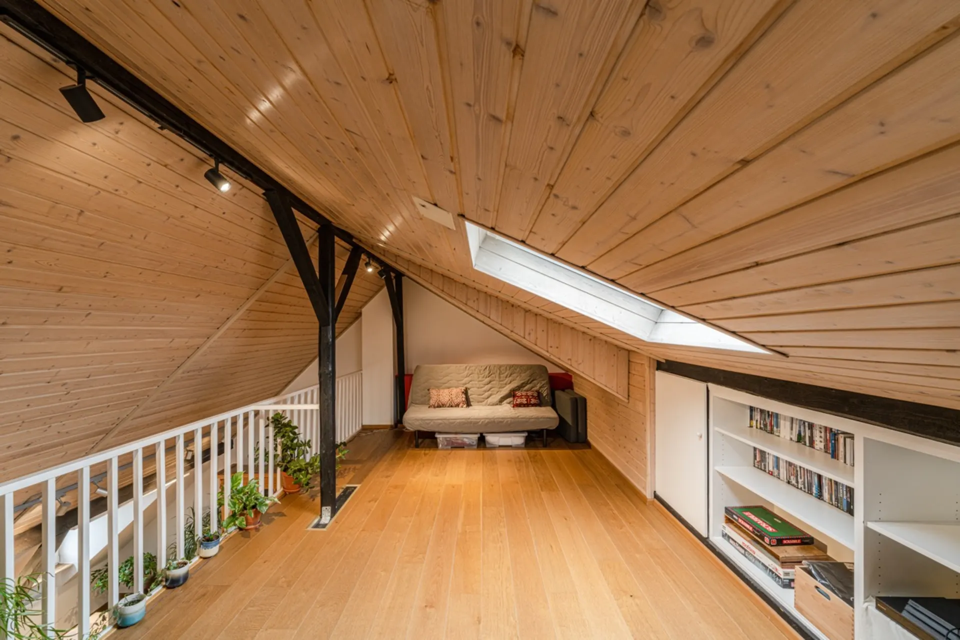 Gemütlicher Dachbodenraum mit Holzdecke, Sofa, Pflanzen, Bücherregal und Dachfenster.
