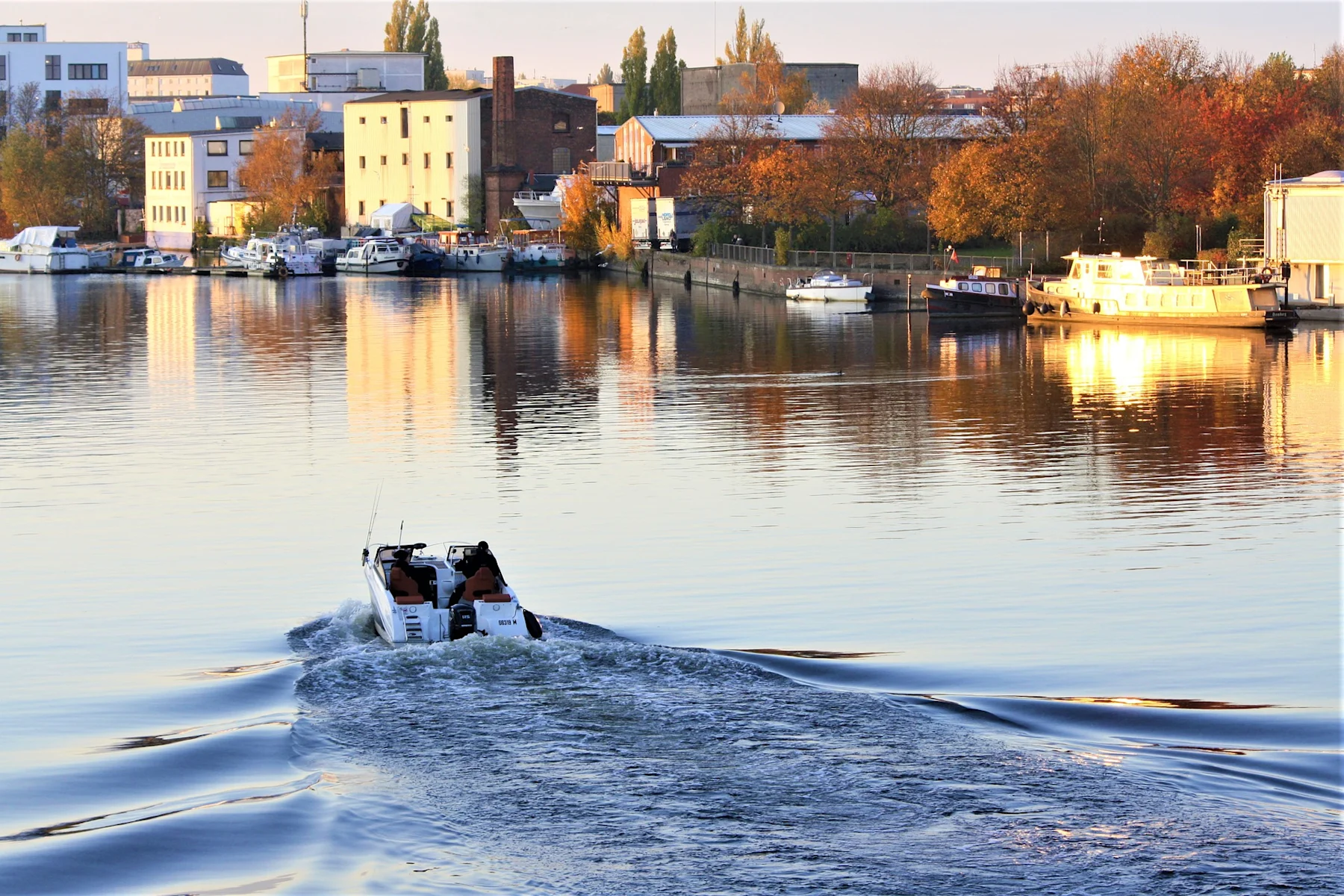 Kleines Motorboot fährt auf einem Fluss, im Hintergrund Ufer mit Gebäuden und Booten.