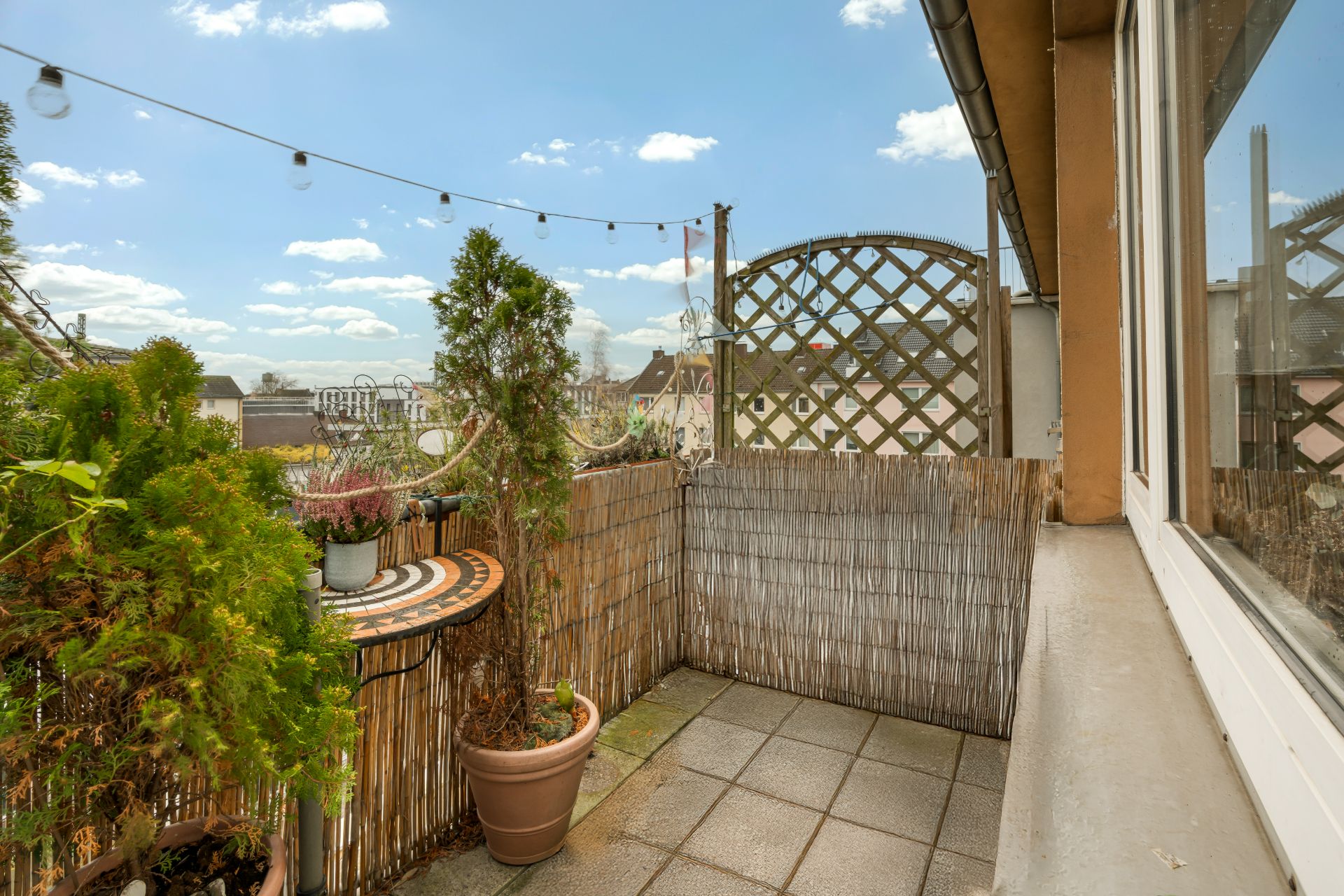 Kleiner Balkon mit Fliesenboden, Pflanzen in Töpfen und Sichtschutz aus Bambus.