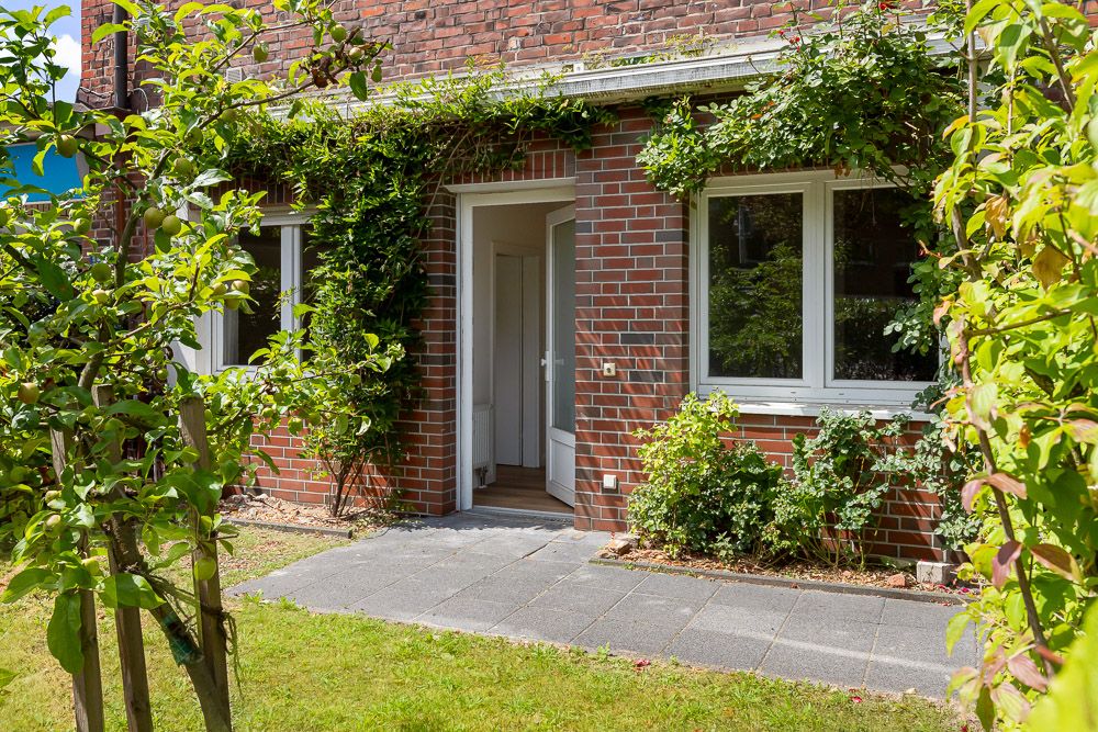 Backsteinfassade mit Tür, zwei Fenstern und Garten mit Pflanzen im Vordergrund.