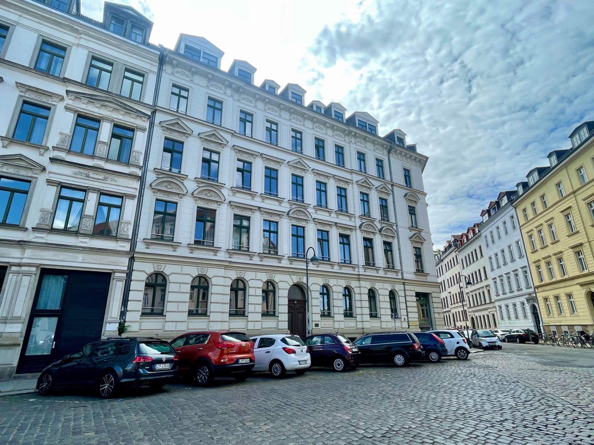 Mehrstöckiges Wohnhaus mit geparkten Autos auf Kopfsteinpflasterstraße.