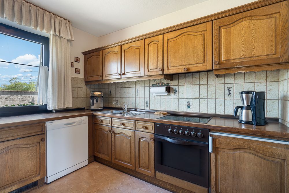 Küche mit Holzschränken, Elektroherd, Geschirrspüler, Spüle, Kaffeemaschine und Fenster.