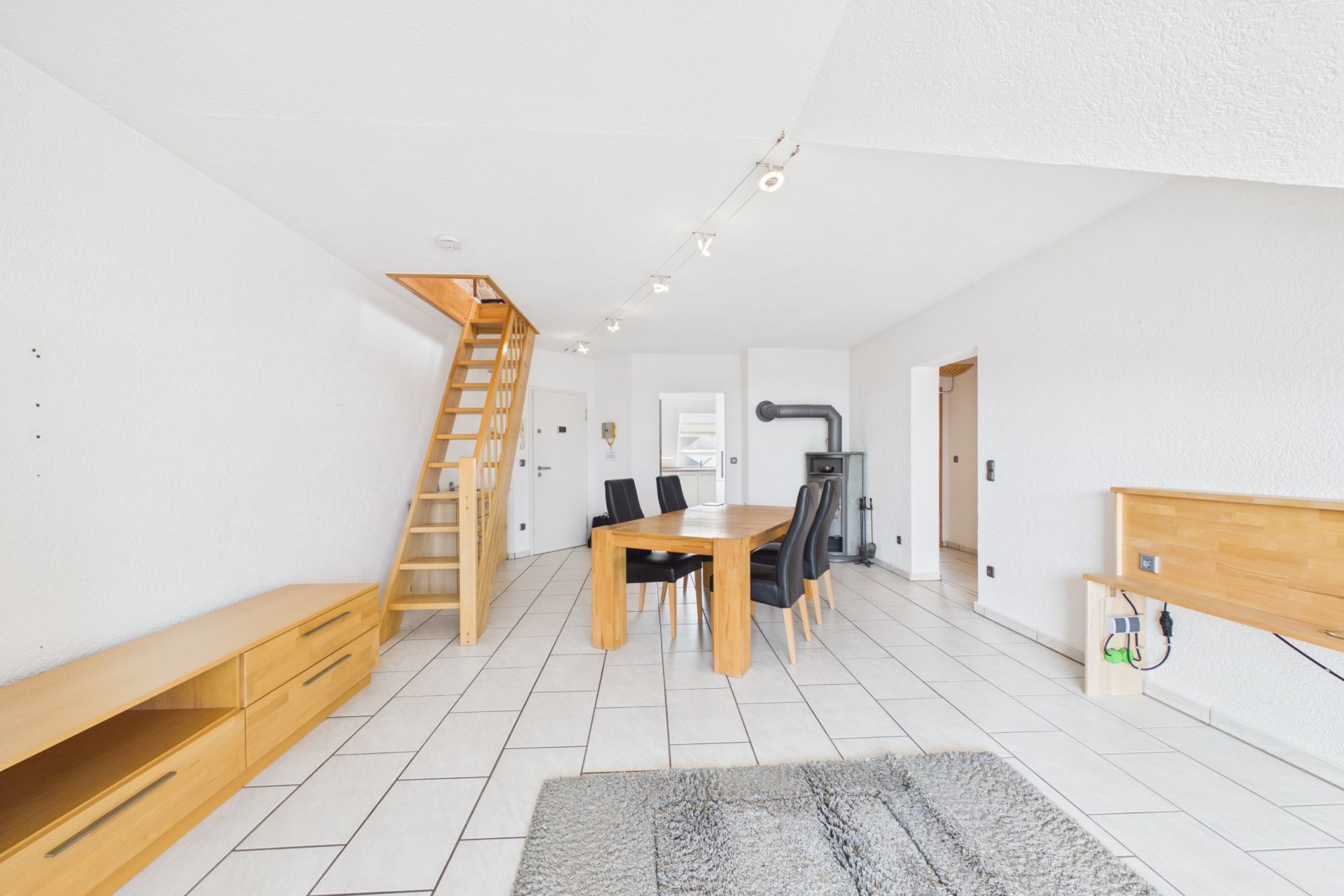 Heller Wohnraum mit Holztisch, schwarzen Stühlen, Holztreppe und Kamin auf weißen Fliesen.