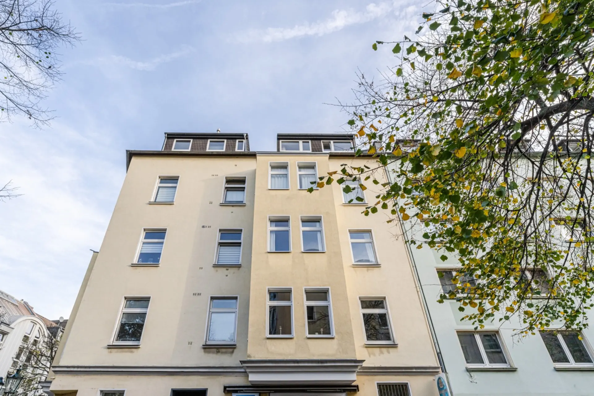 Mehrstöckiges Wohnhaus mit gelber Fassade und mehreren Fenstern, Baum rechts im Vordergrund.