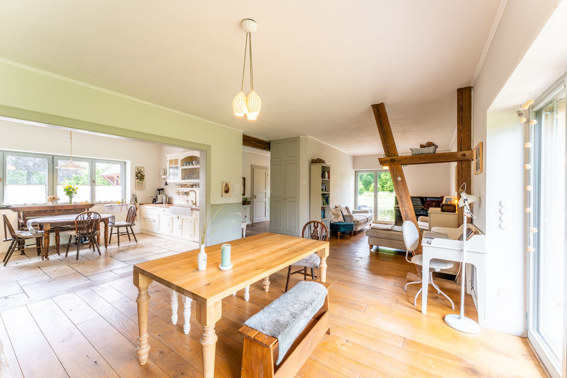 Helles Wohnzimmer mit Holzboden, Esstisch, Küche und großen Fenstern mit Blick nach draußen.