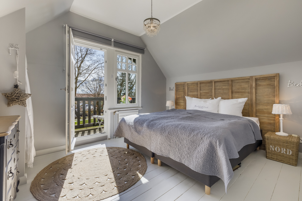 Helles Schlafzimmer mit Doppelbett, grauer Tagesdecke und offenem Zugang zum Balkon.