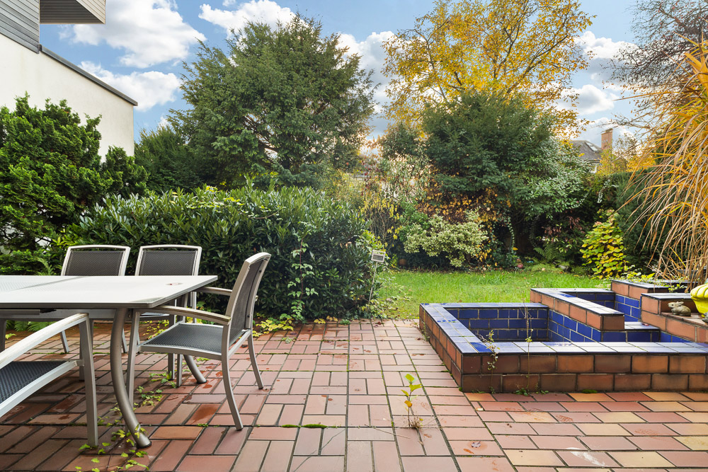 Gepflasterte Terrasse mit Gartentisch, Stühlen und kleinem Brunnen, umgeben von grünen Sträuchern und Bäumen.