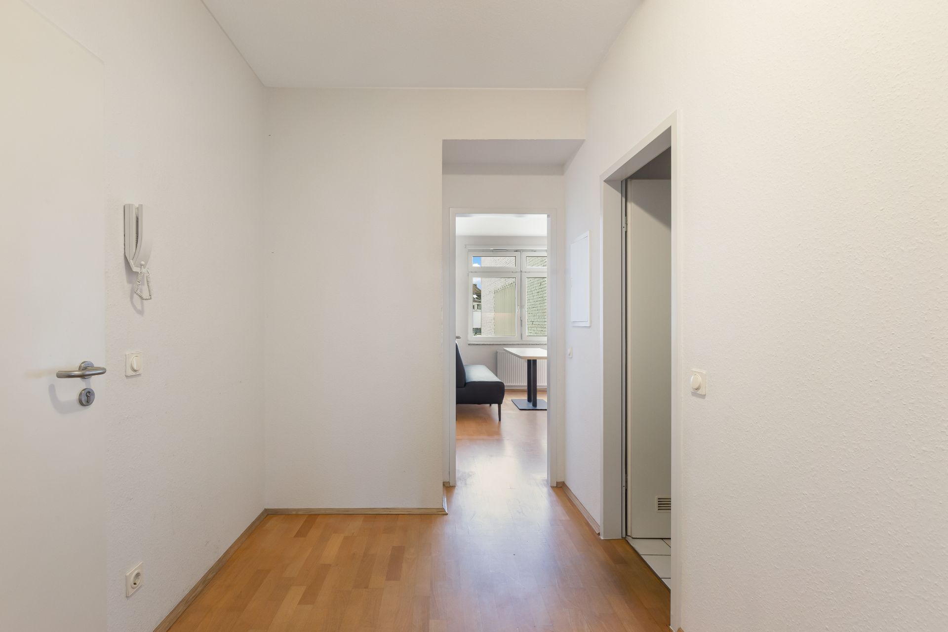 Heller Flur mit Holzboden, weißen Wänden und Sicht auf ein Wohnzimmer im Hintergrund.