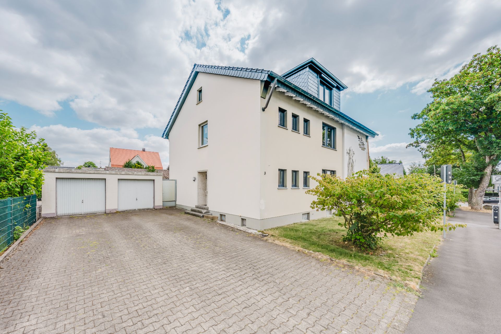 Zweistöckiges Wohnhaus mit Garagen und gepflasterter Einfahrt, umgeben von Bäumen.