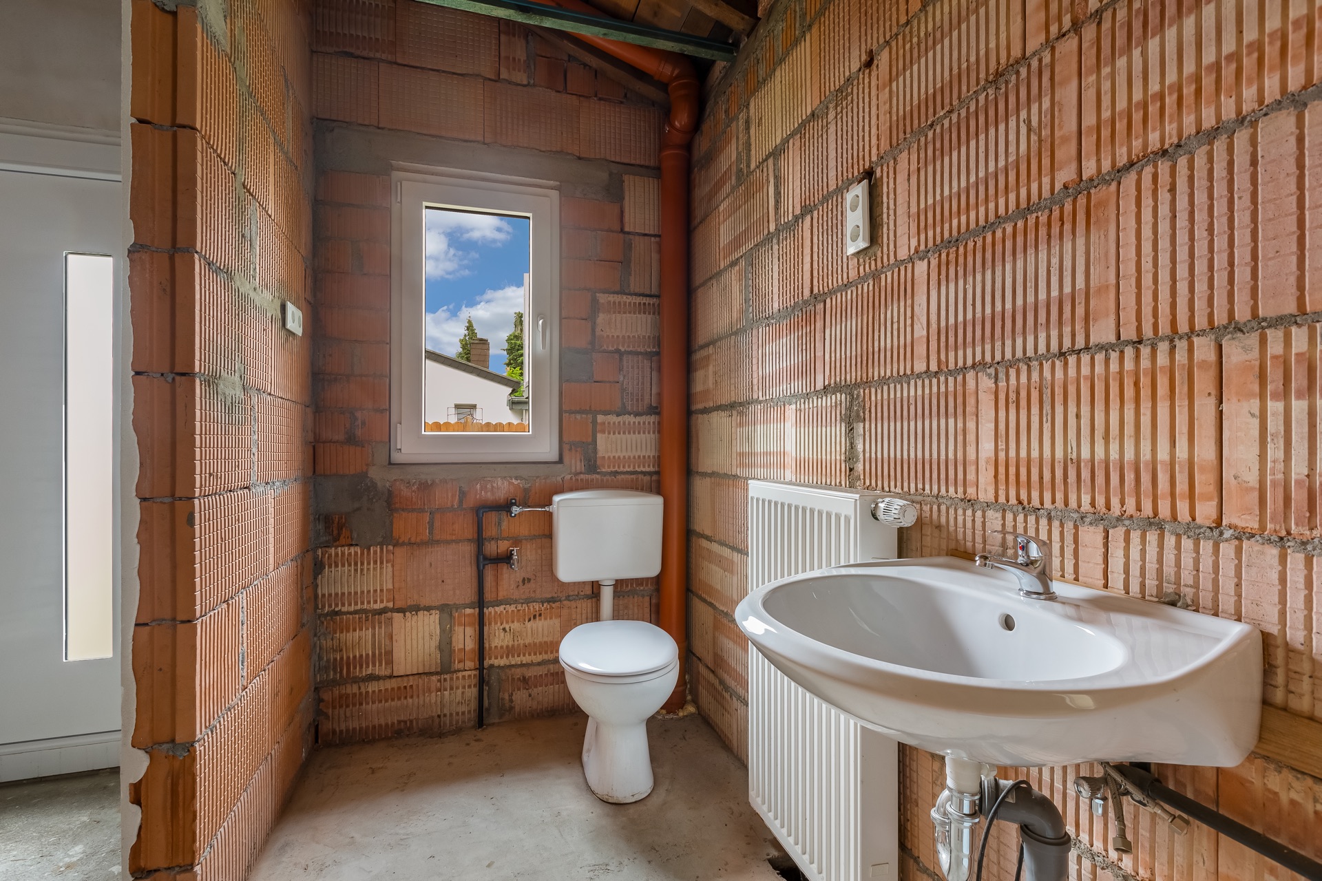 Unfertiges Badezimmer mit Toilette, Waschbecken, Fenster und sichtbaren Ziegelwänden.