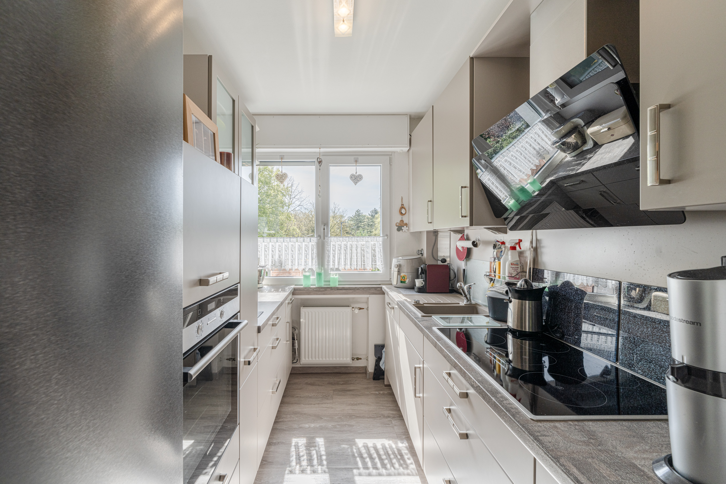 Schmale, moderne Küche mit weißen Schränken, Elektroherd, Spüle und Fenster mit Ausblick.