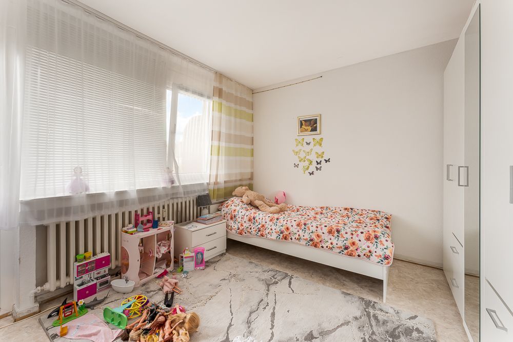 Helles Kinderzimmer mit Einzelbett, Spielzeug auf dem Boden und großem Fenster mit Gardinen.