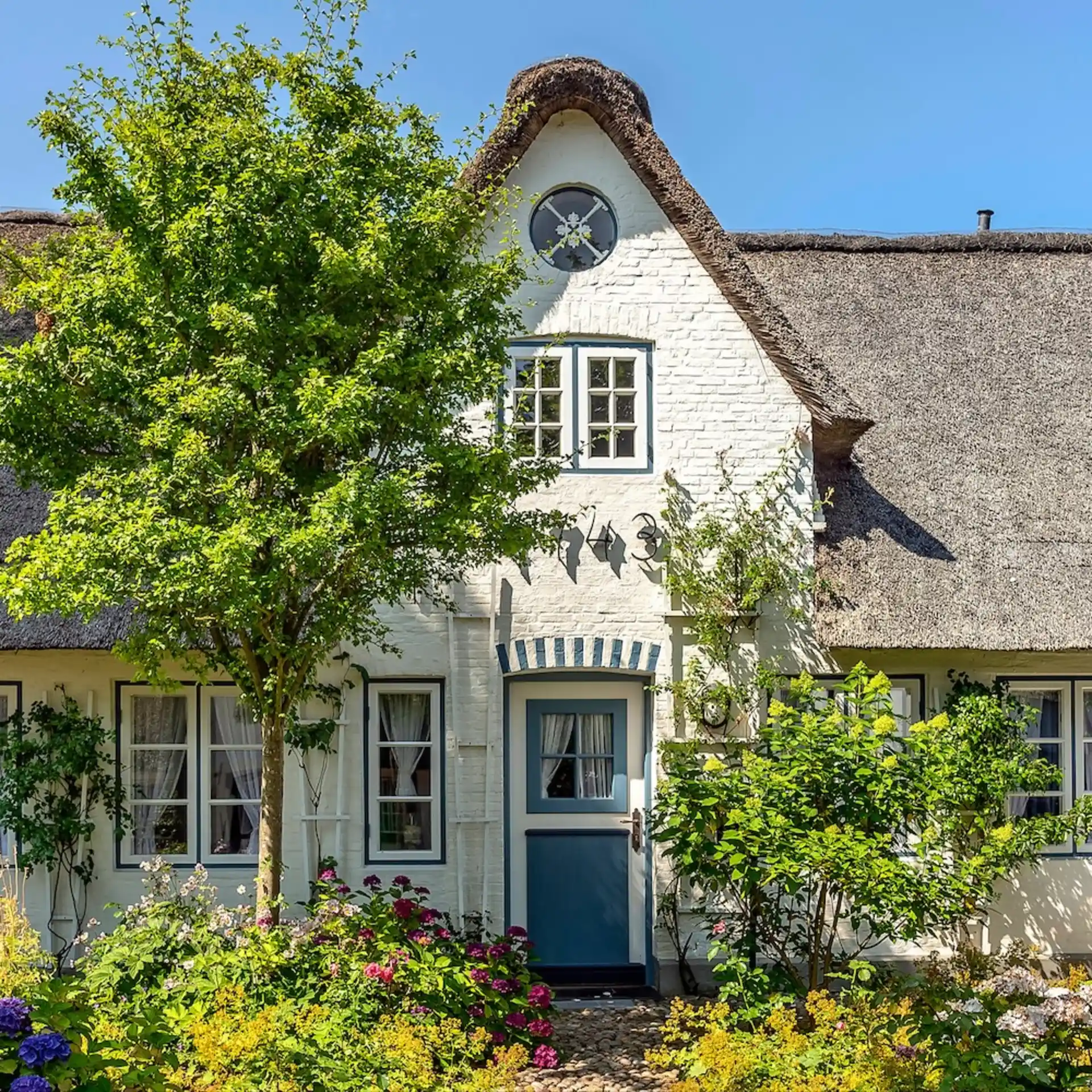 Gemütliches weißes Haus mit Reetdach, blauer Tür und blühendem Garten im Vordergrund.