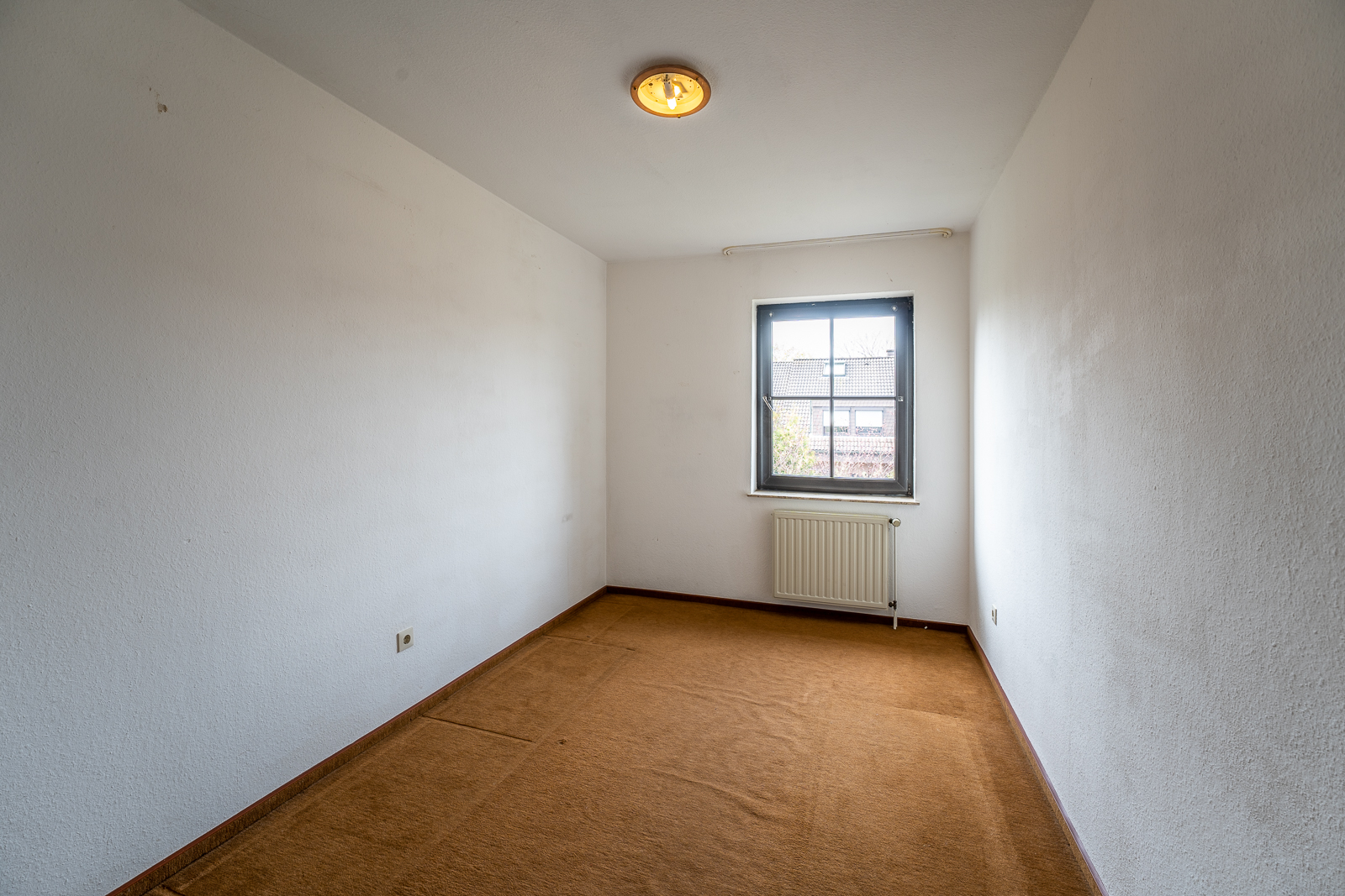 Leeres Zimmer mit braunem Teppich, weiß gestrichenen Wänden und einem Fenster mit Heizkörper.