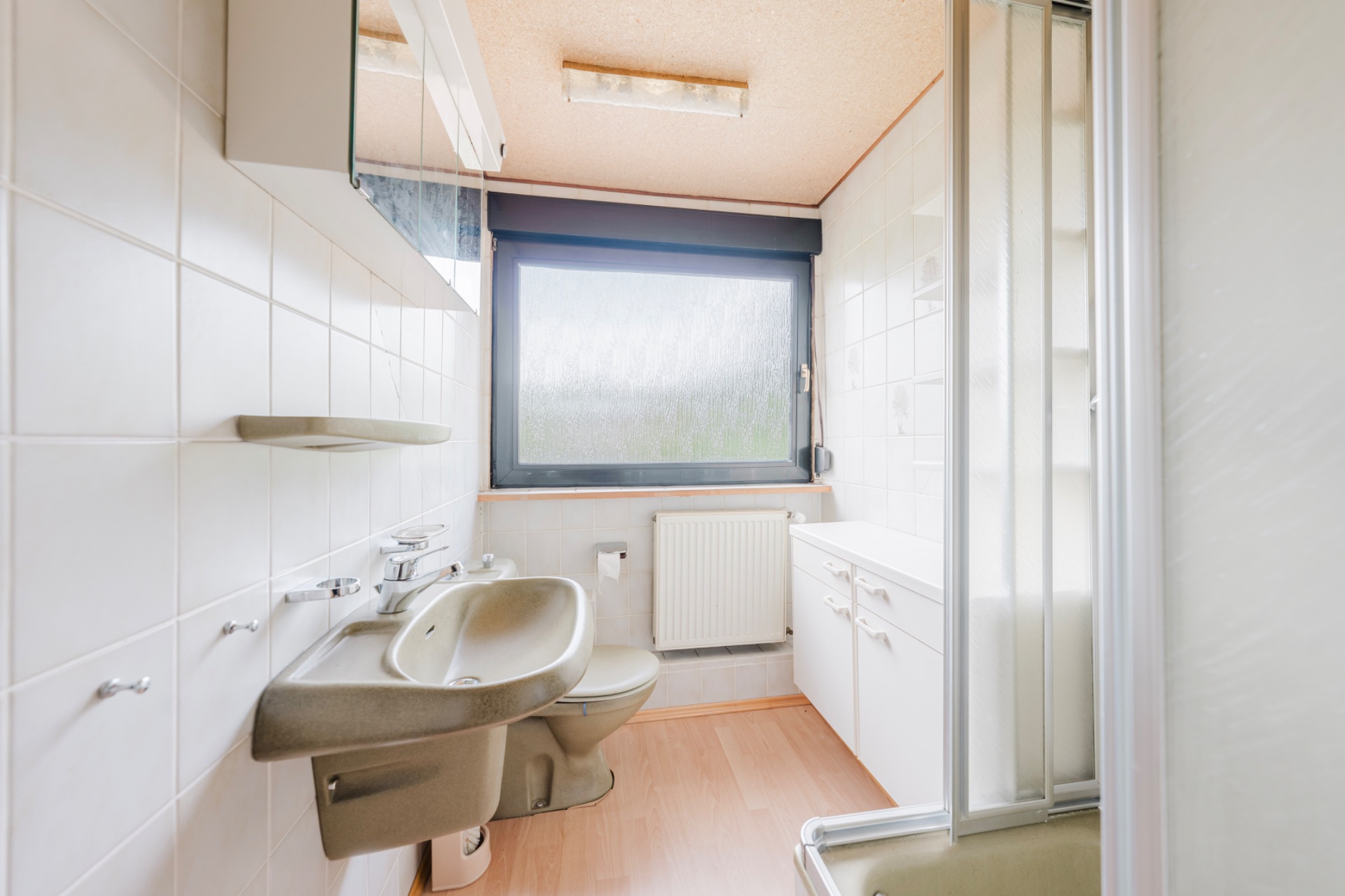 Helles Badezimmer mit Waschbecken, Toilette, Dusche, großem Fenster und weißen Fliesen.