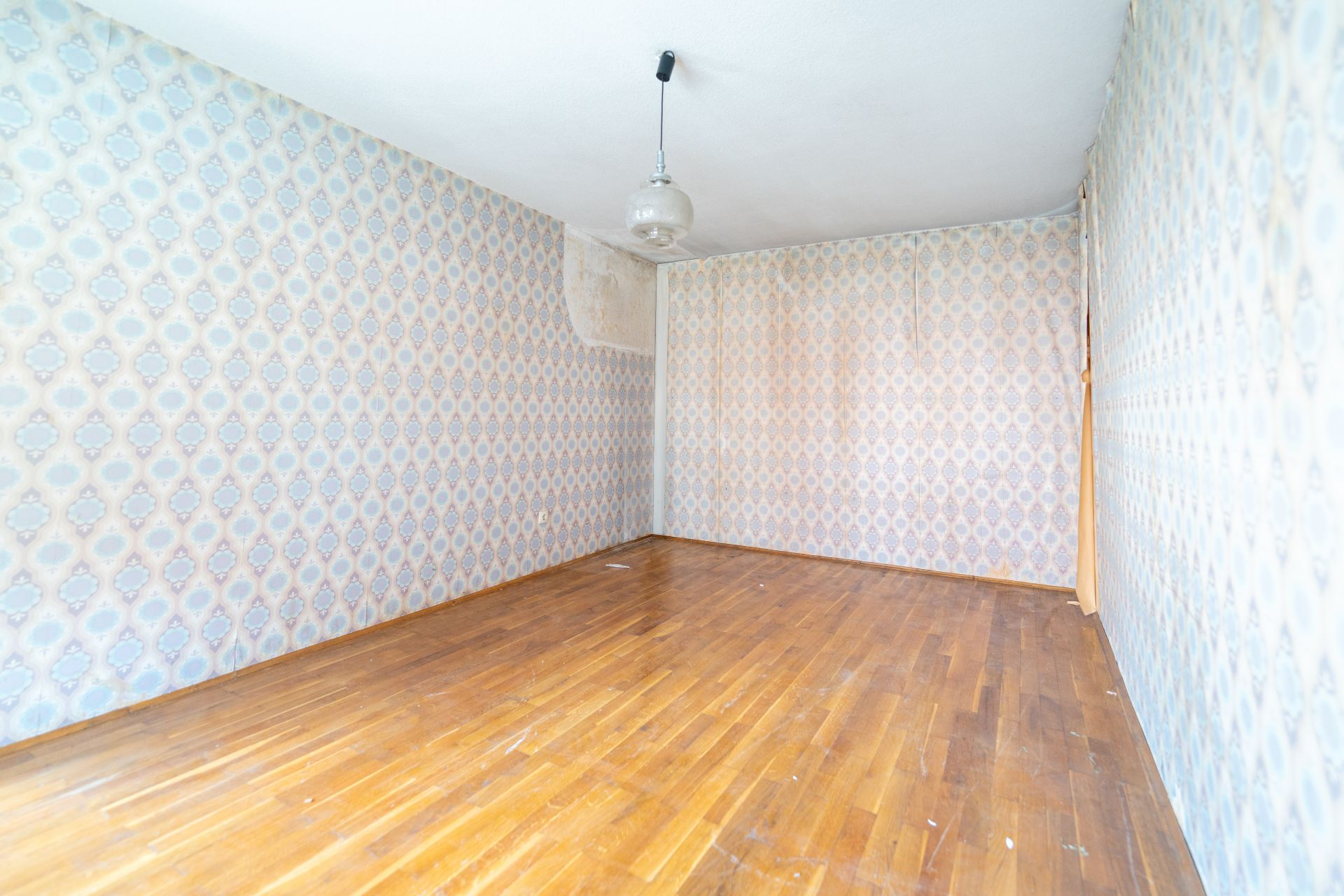 Leerer Raum mit gemusterten Tapeten und Holzfußboden, einzelne Deckenlampe.