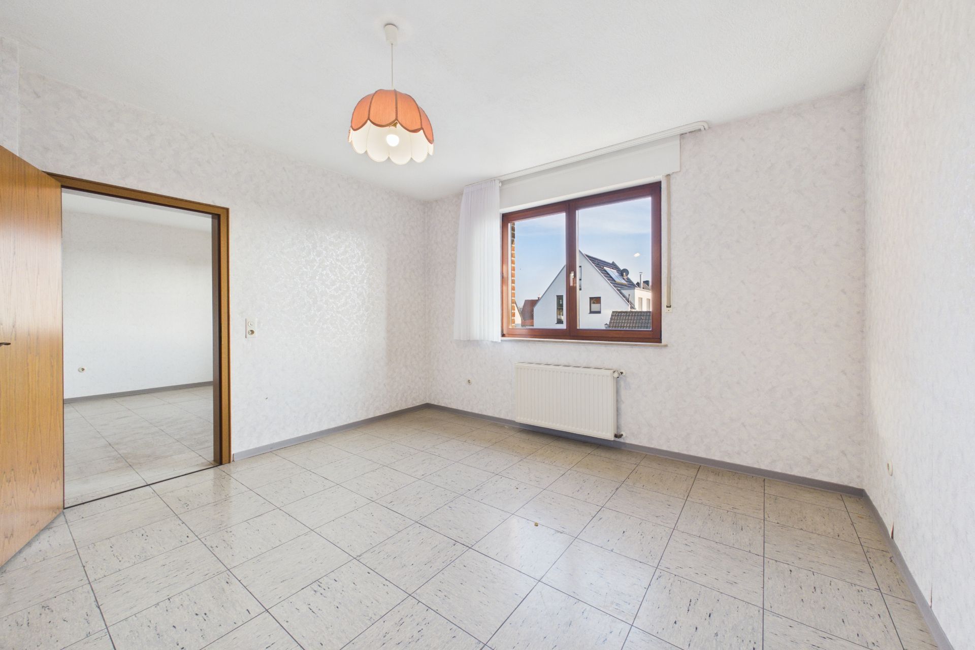 Leeres, helles Zimmer mit weißer Tapete, Fliesenboden und großem Fenster mit Blick nach draußen.