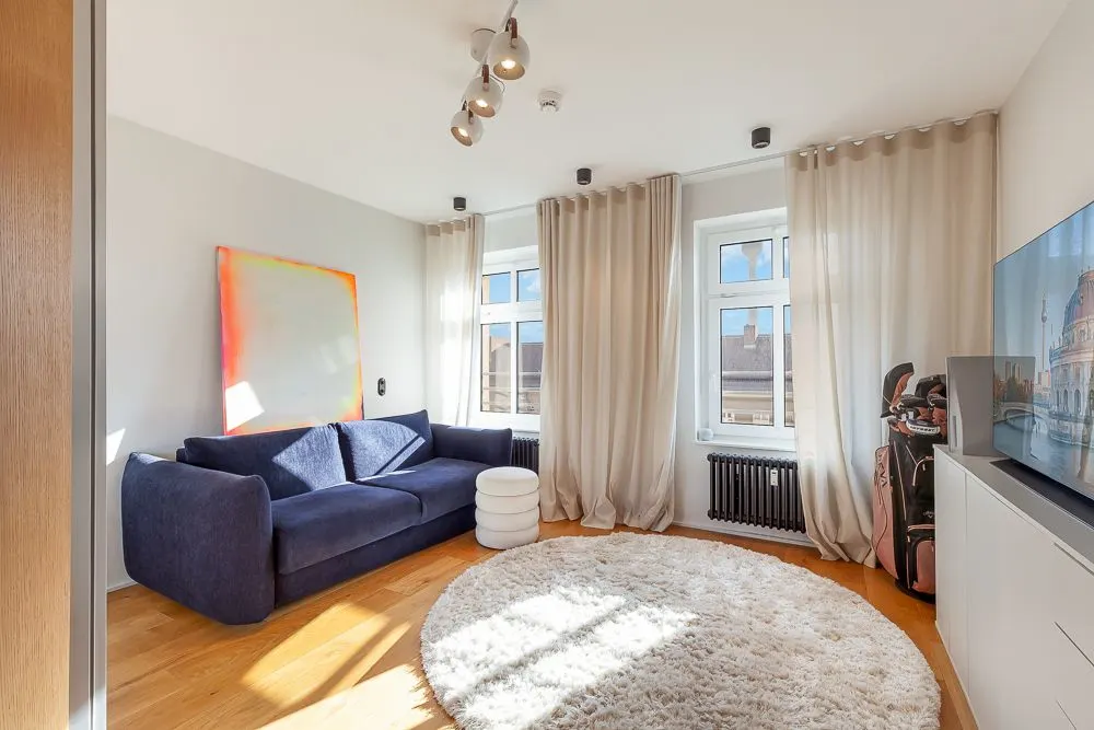 Helles Wohnzimmer mit blauem Sofa, rundem Teppich, zwei Fenstern und TV an der Wand.