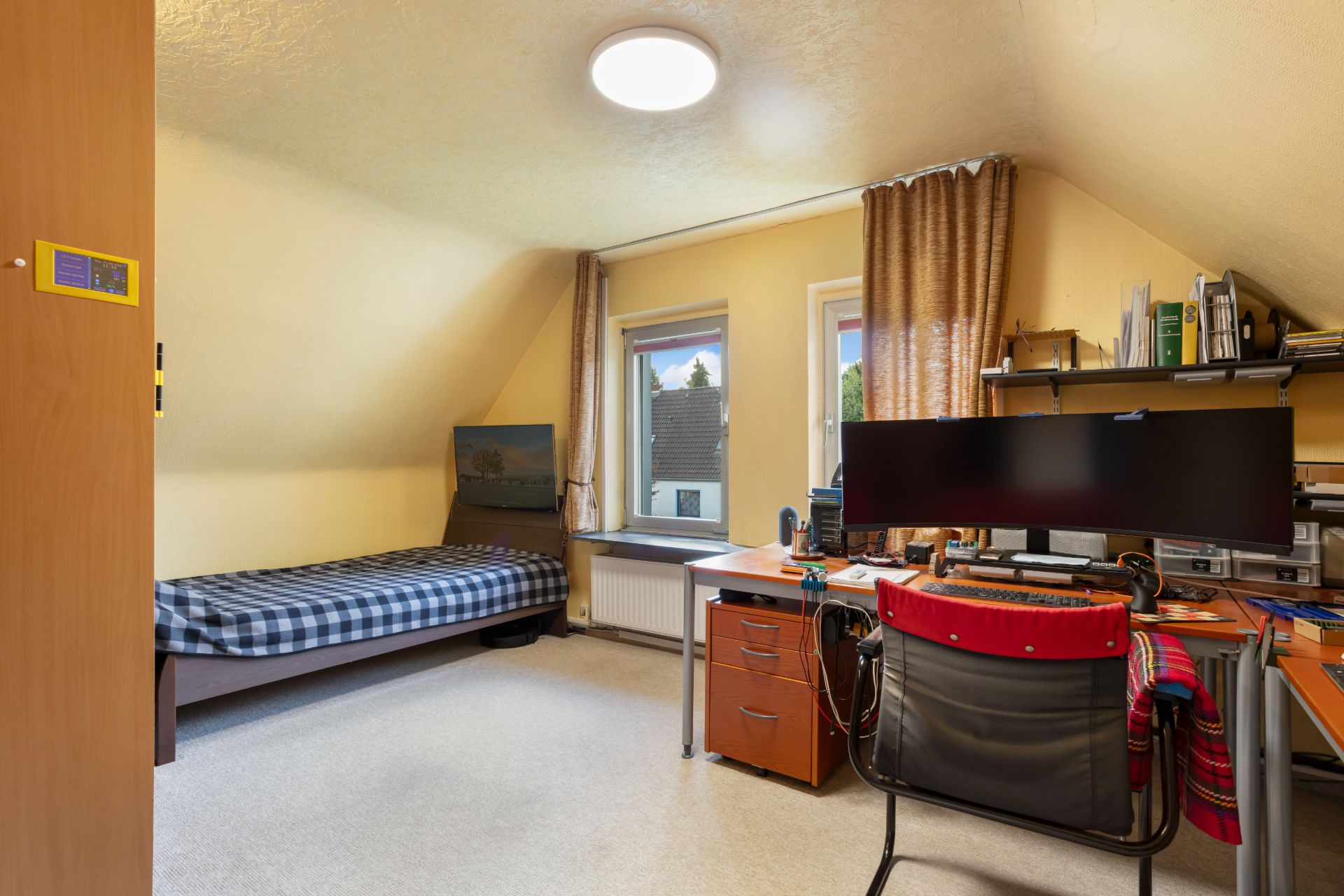 Helles Zimmer mit Bett, Schreibtisch, Stuhl und großem Monitor, zwei Fenstern und Regalen an der Wand.