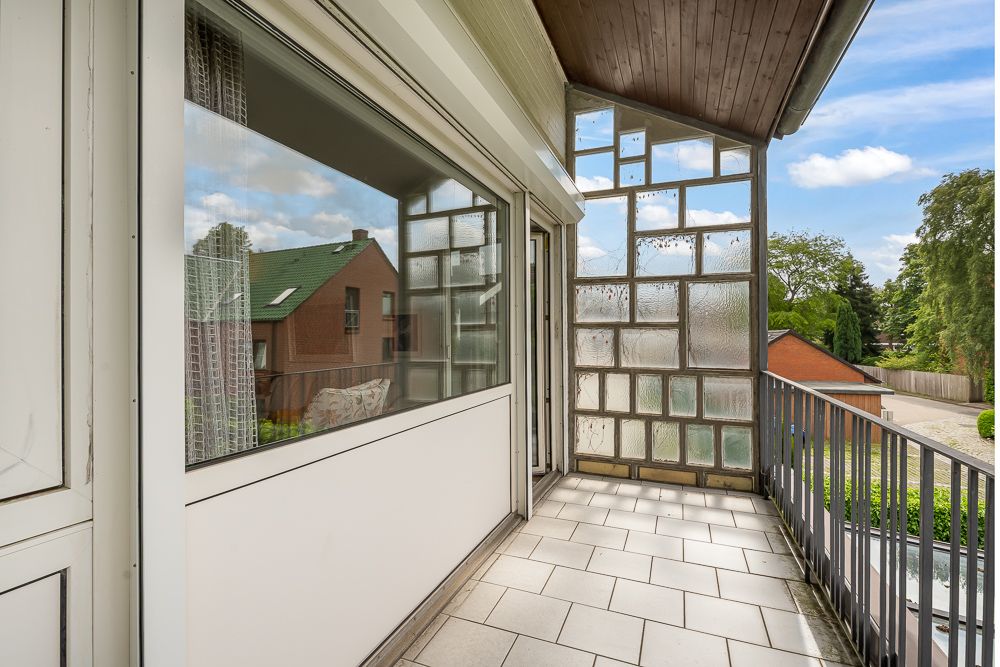 Überdachter Balkon mit Fliesenboden und teilweiser Glaswand, Ausblick auf Nachbarhäuser und Bäume.