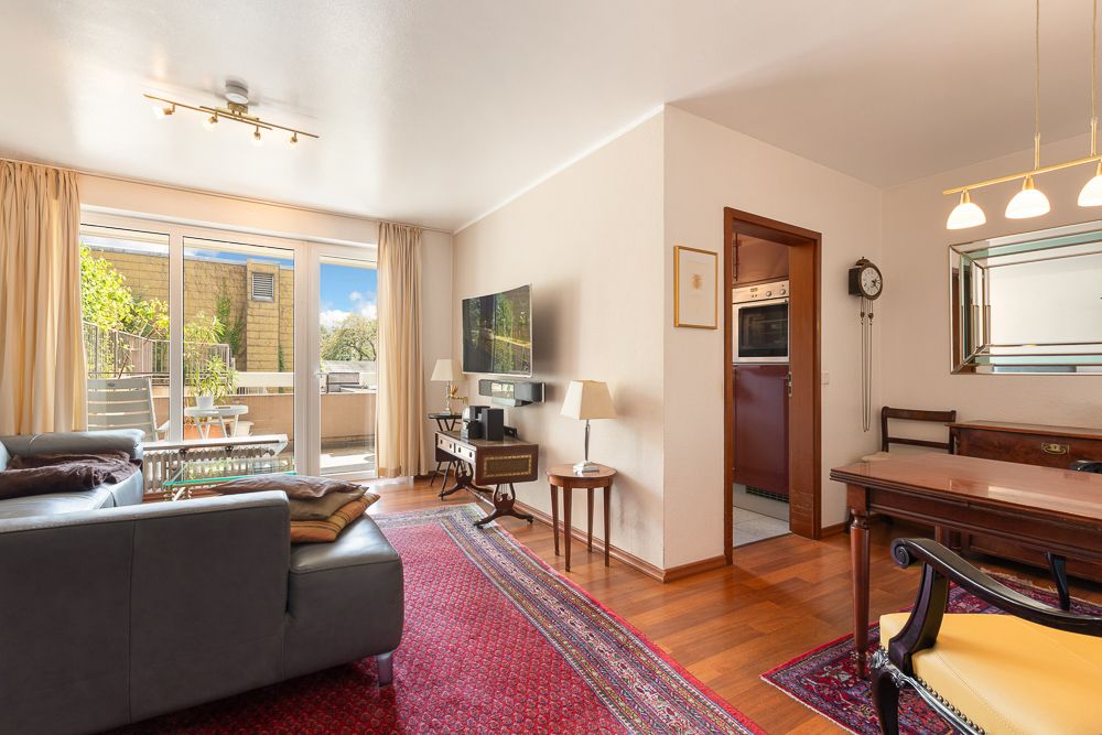 Helles Wohnzimmer mit Sofa, Teppich, Esstisch und Zugang zu einem Balkon mit Pflanzen.