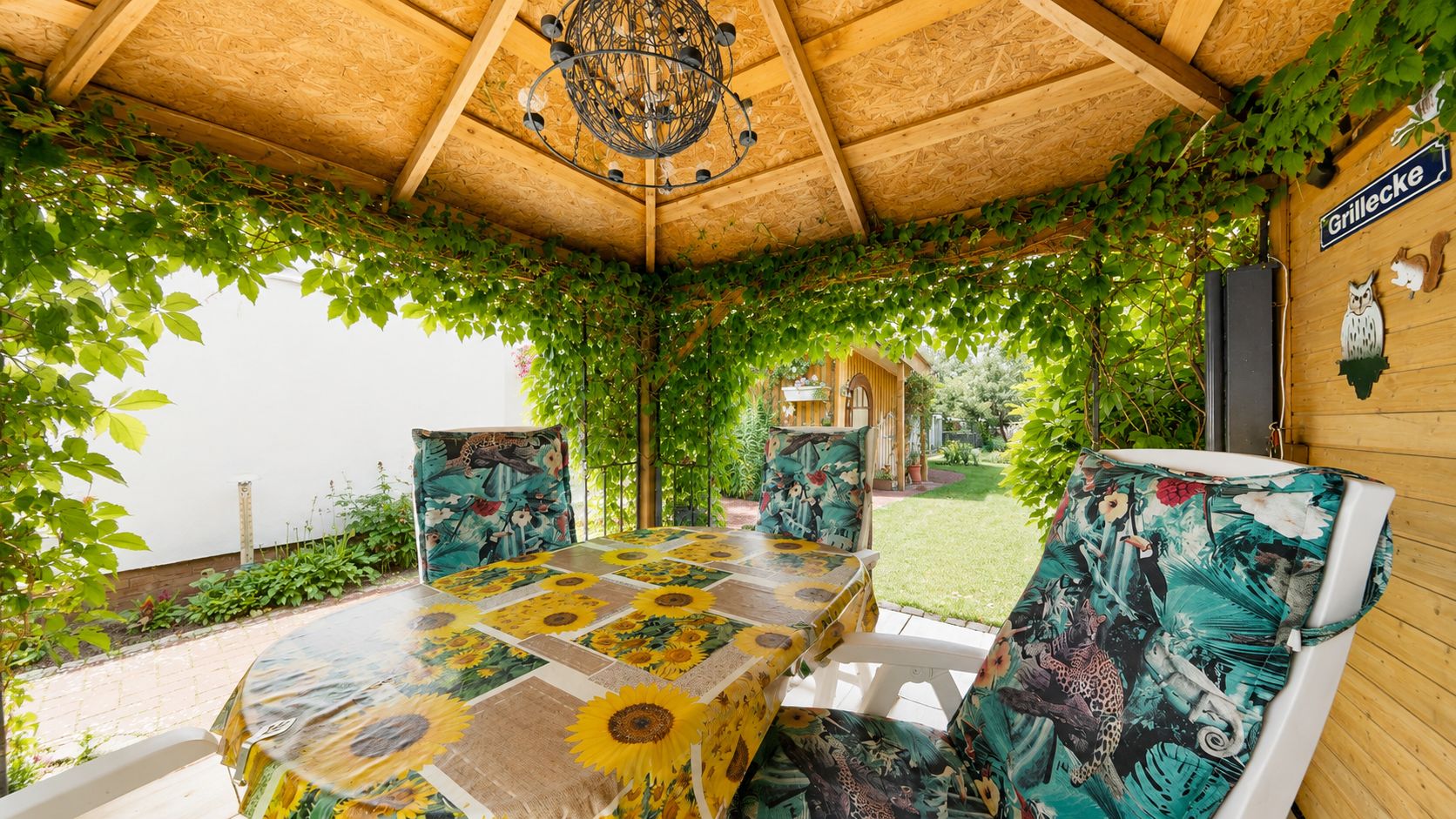 Überdachte Terrasse mit Tisch und gepolsterten Stühlen, umgeben von Efeu und Blick in den Garten.
