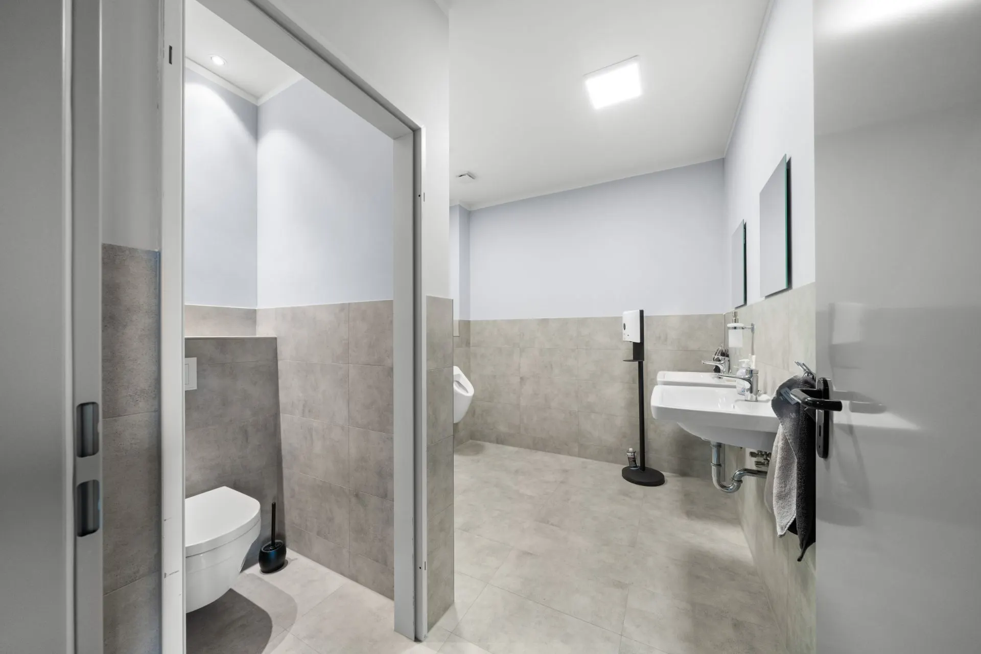 Modernes Badezimmer mit Toilette, Waschbecken, Spiegel und Urinal, helle Fliesen und weiße Wände.