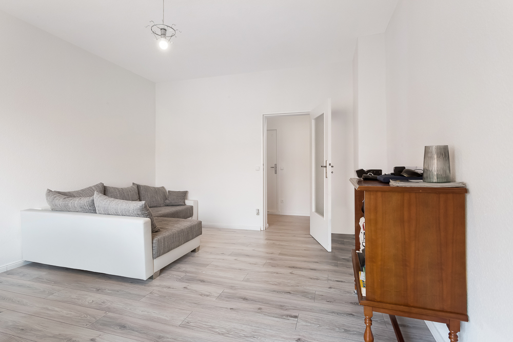 Helles Wohnzimmer mit grauem Sofa, Holzkommode und offen stehender Tür auf hellem Holzboden.