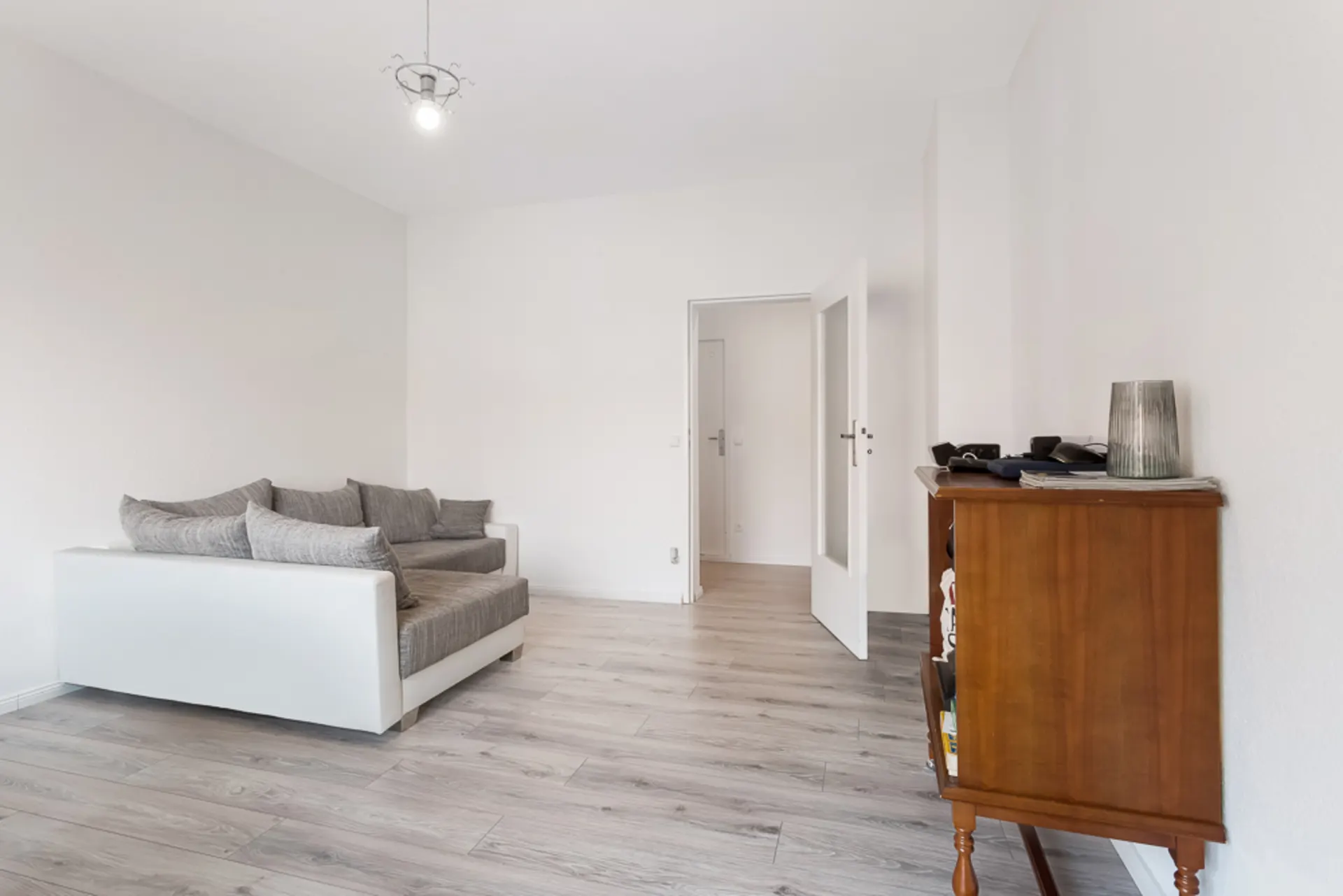 Helles Wohnzimmer mit grauem Sofa, Holzkommode und offen stehender Tür auf hellem Holzboden.