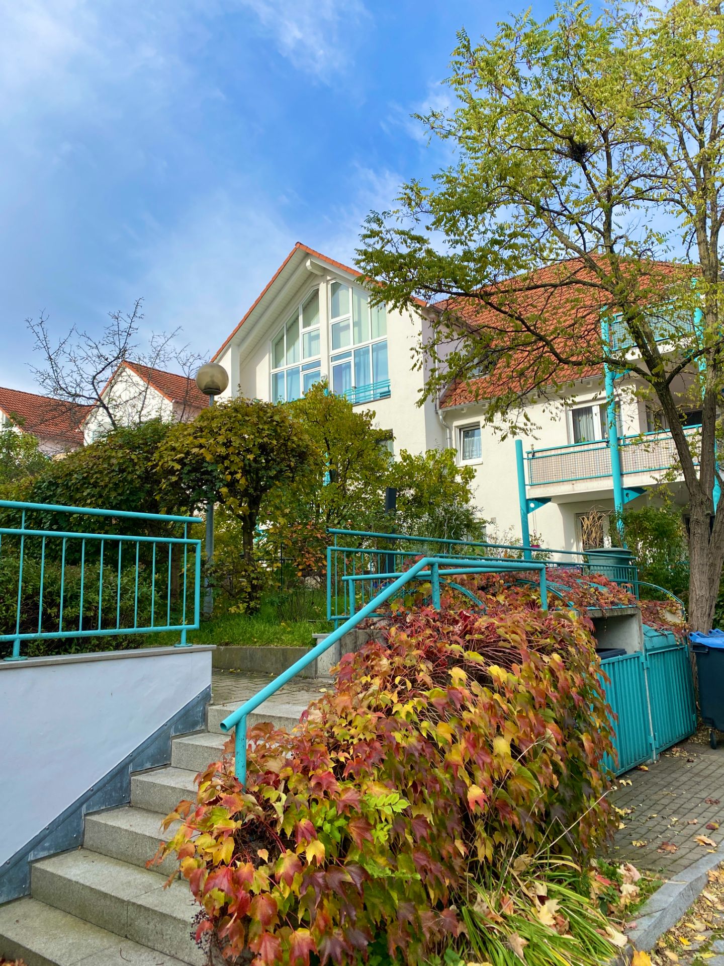 Außenansicht eines Wohnhauses mit rotem Dach, Treppe, blauem Geländer und herbstlicher Bepflanzung.