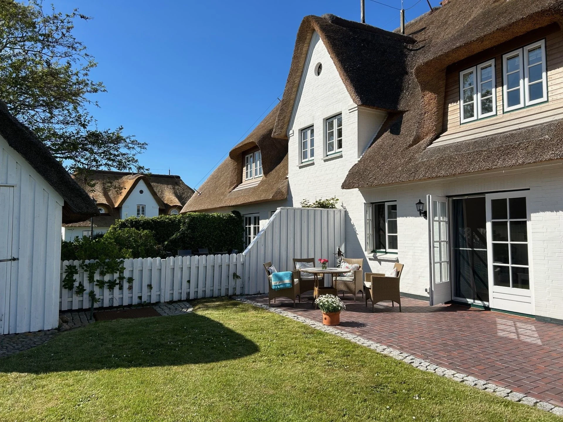 Gepflegter Garten mit Terrasse, Sitzgruppe und weißem Zaun vor einem Haus mit Reetdach.