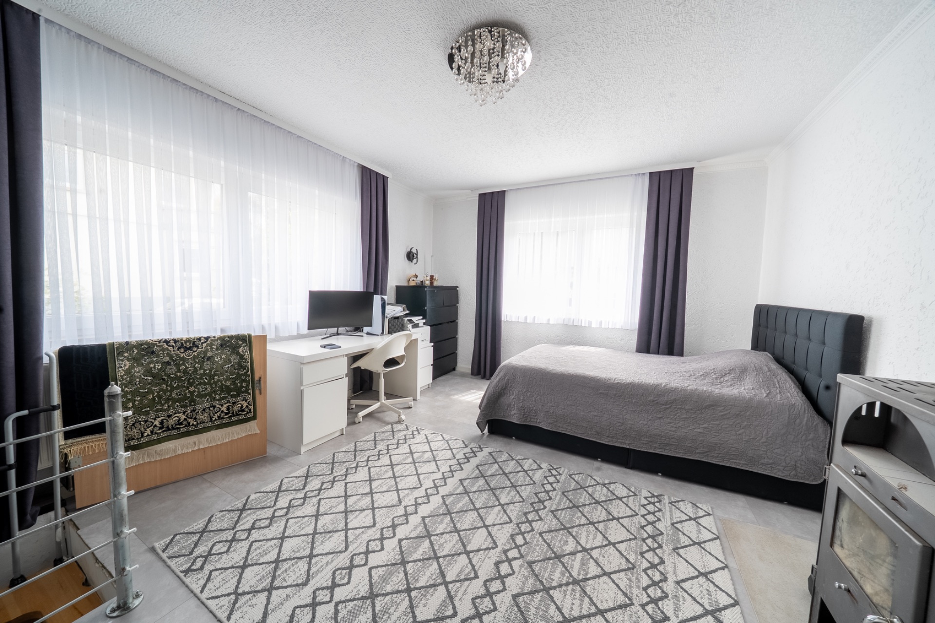 Helles Schlafzimmer mit Bett, Teppich, Schreibtisch, Stuhl und zwei Fenstern mit Vorhängen.