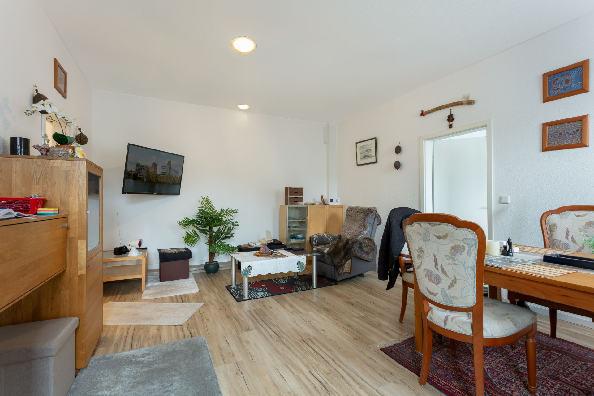 Wohnzimmer mit Holzboden, TV an der Wand, Esstisch, Sessel und Zimmerpflanze.