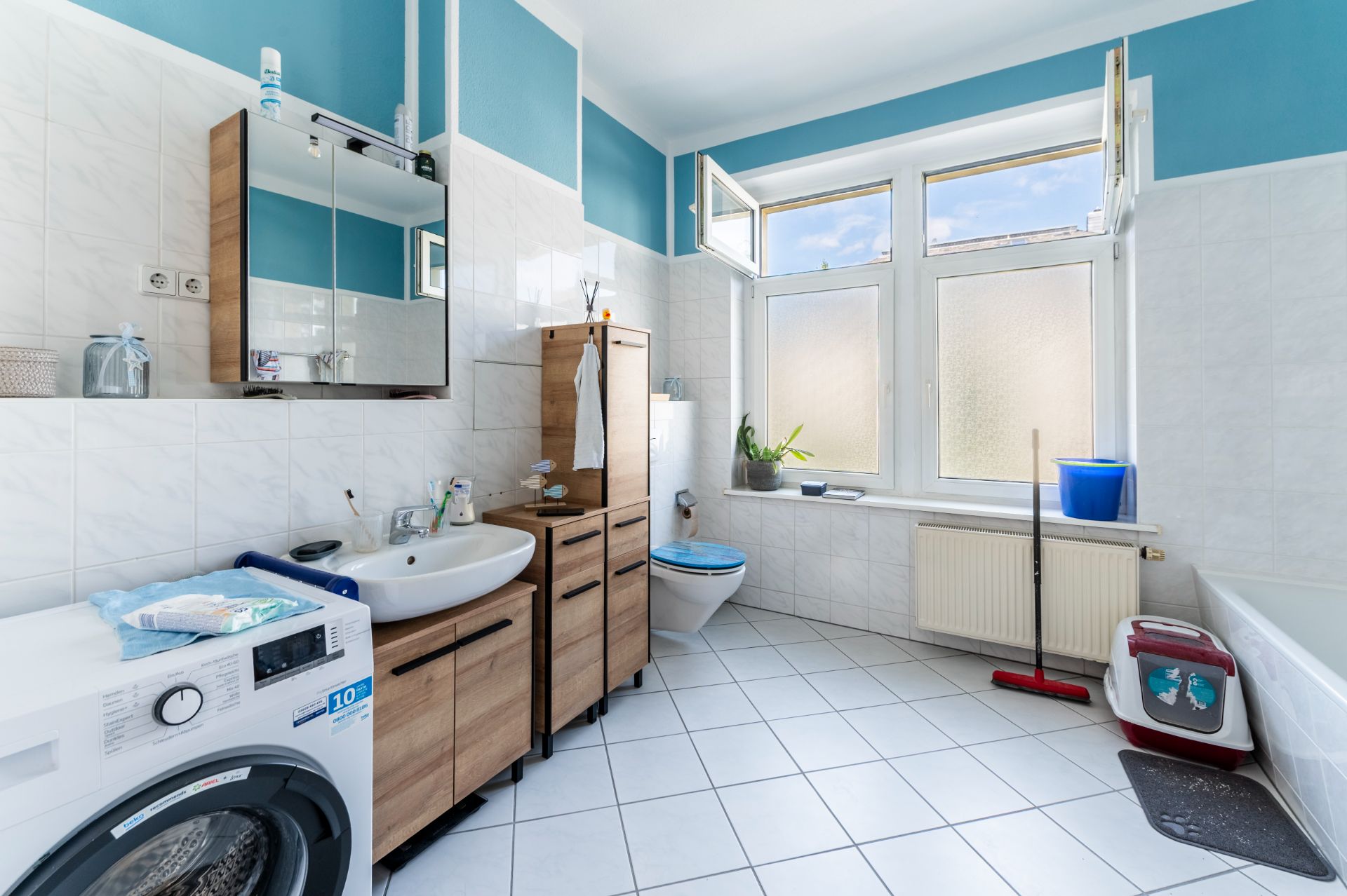 Modernes Badezimmer mit Waschmaschine, Holzschränken, Waschbecken, WC, Badewanne und großen Fenstern.