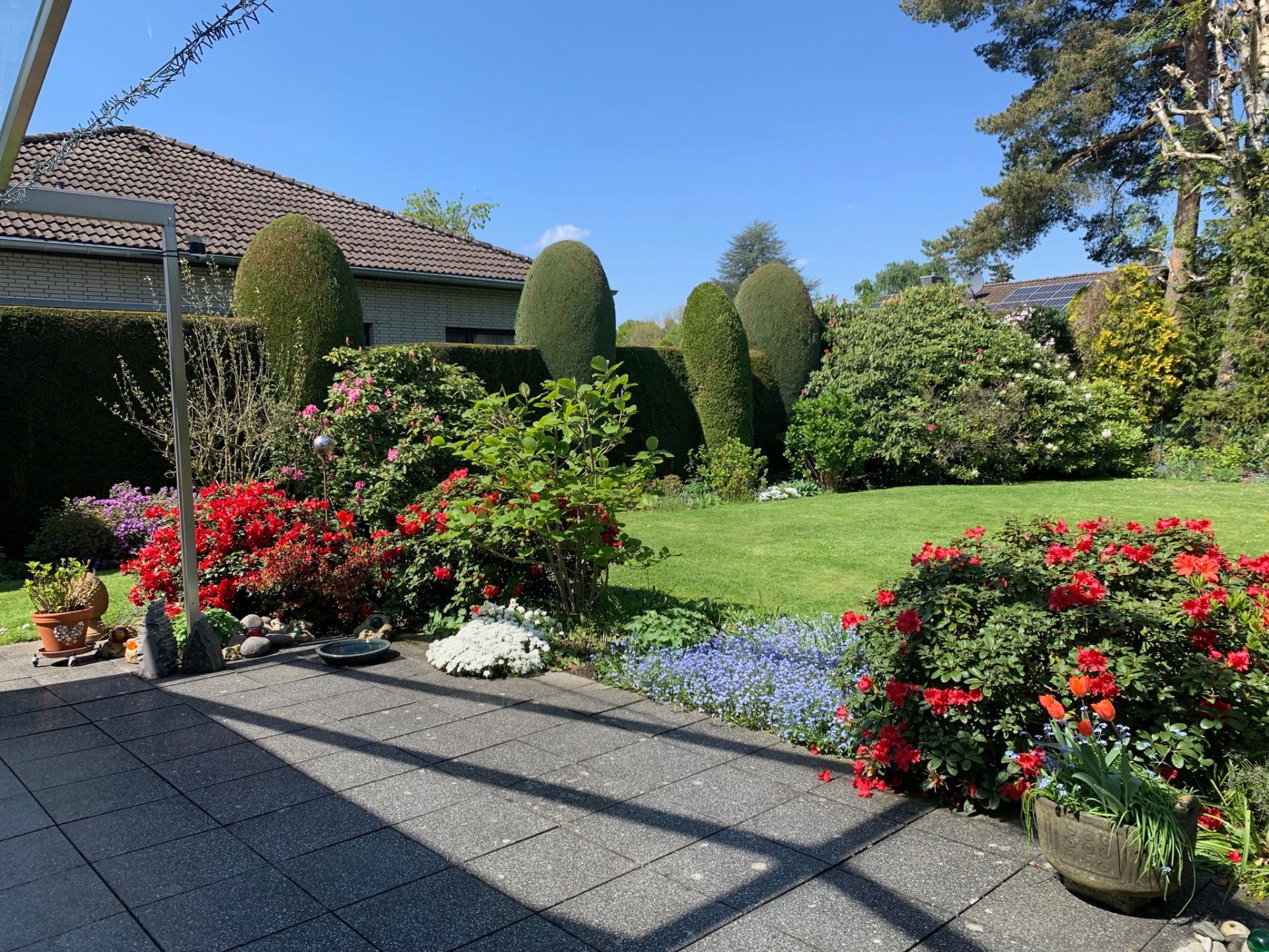 Gepflegter Garten mit Blumen, Rasenfläche und Terrasse, umgeben von Sträuchern und Bäumen.