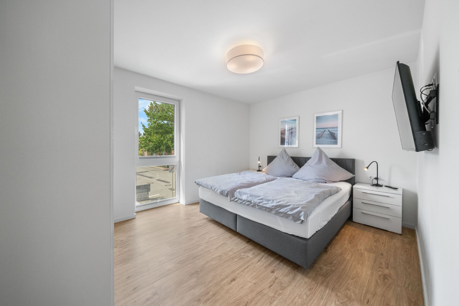 Helles Schlafzimmer mit Doppelbett, Nachttischen, Wandbildern, Fernseher und großem Fenster.