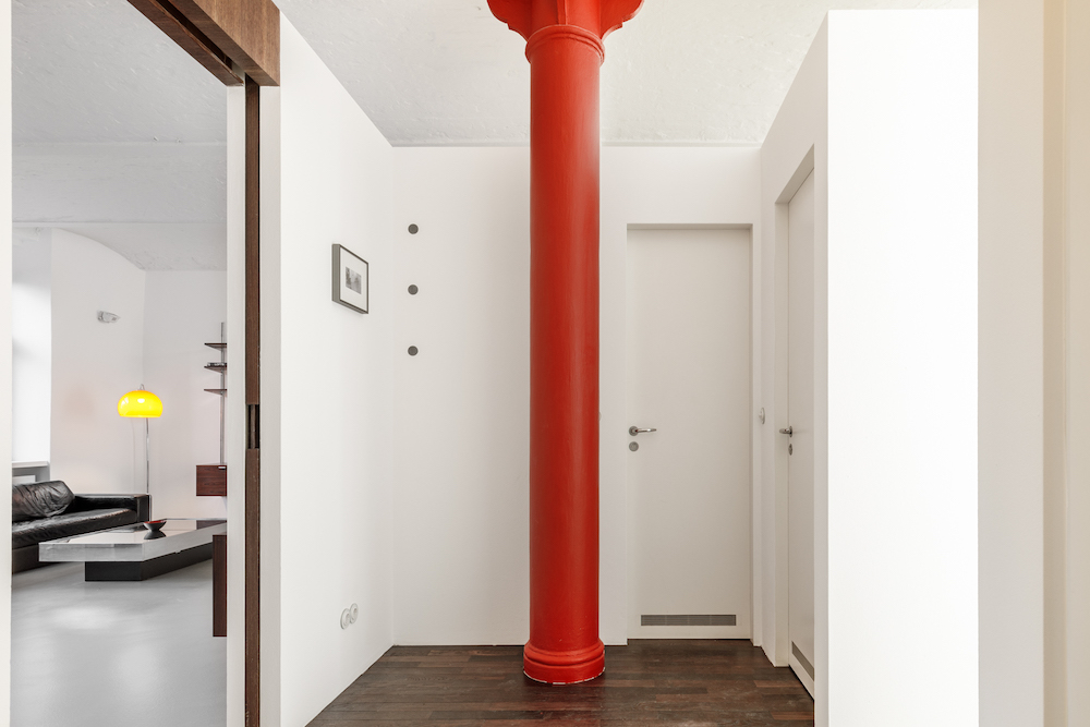 Flur mit roter Säule, weißer Wand und zwei weißen Türen, Holzboden.