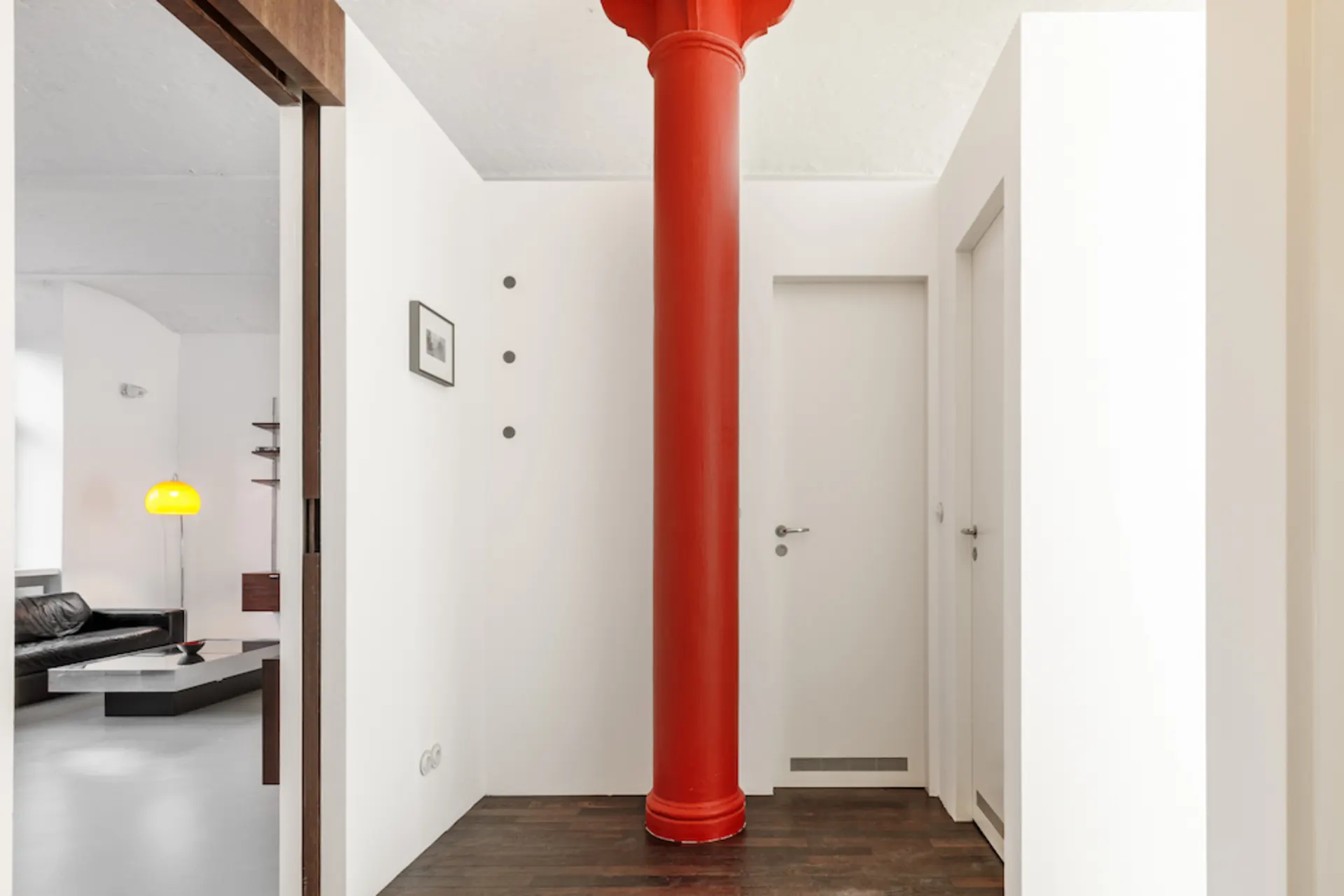 Flur mit roter Säule, weißer Wand und zwei weißen Türen, Holzboden.