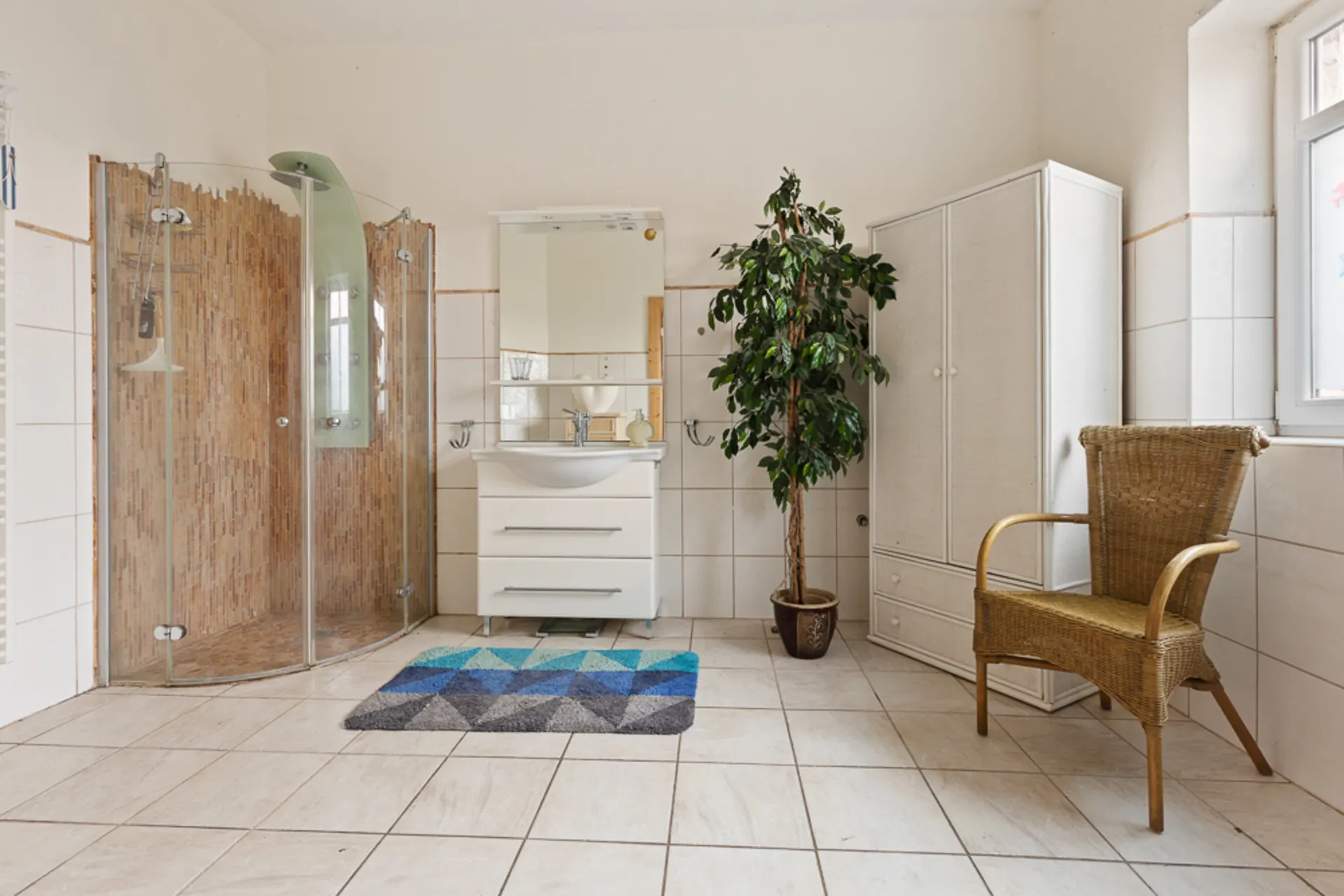 Helles Badezimmer mit Dusche, Waschbecken, Schrank, Pflanze und geflochtenem Stuhl.