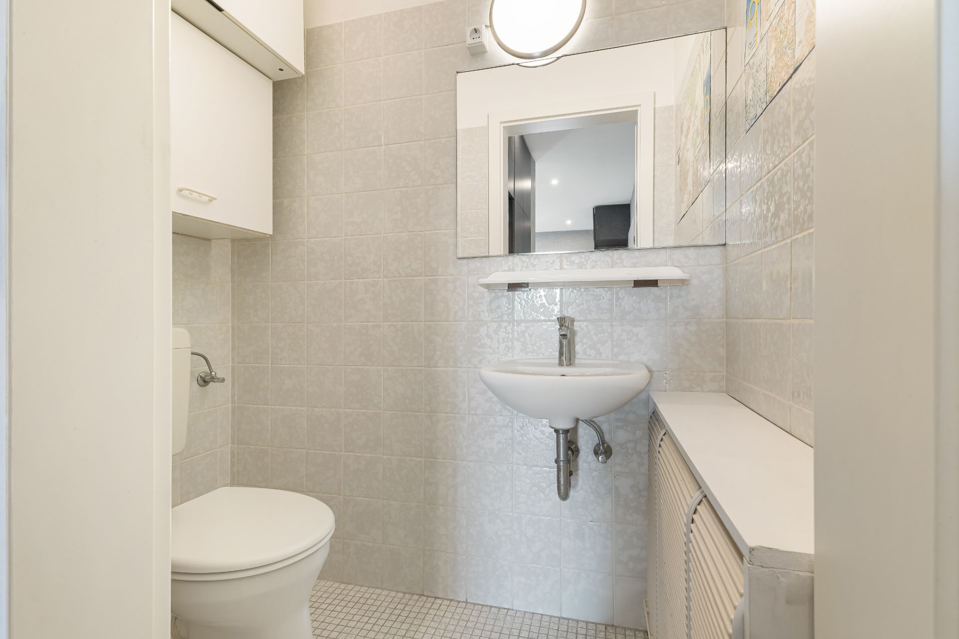 Kleines Badezimmer mit Toilette, Waschbecken, Spiegel und Hängeschrank.